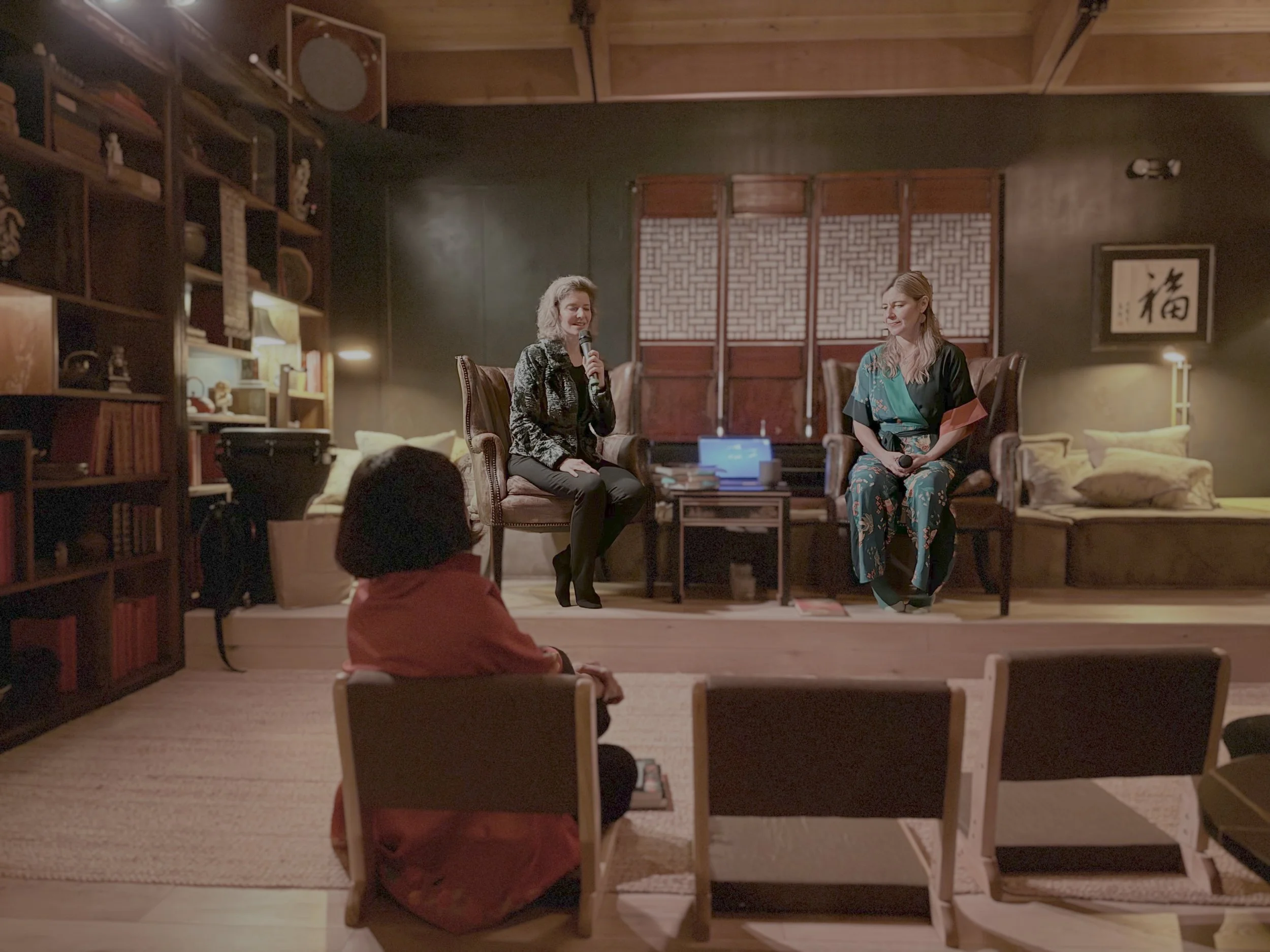 Two women are seated on a stage having a conversation, each holding a microphone. One woman has blonde hair and wears a black jacket, the other has light brown hair and wears a traditional Japanese kimono. An audience member sits in the foreground, f