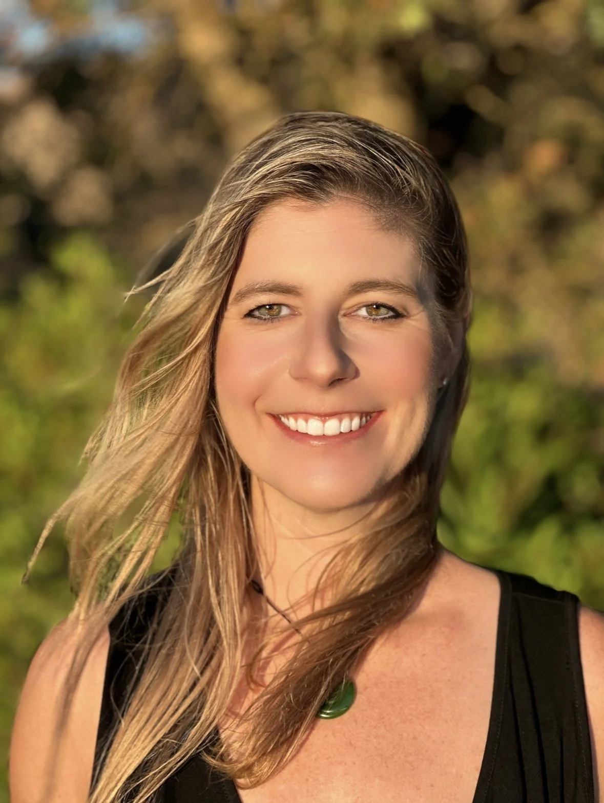 A woman (Caitlin Krause) keynote speaker, with long, light brown hair smiling outdoors, wearing a black sleeveless top and a green necklace, with a background of blurred greenery and trees.