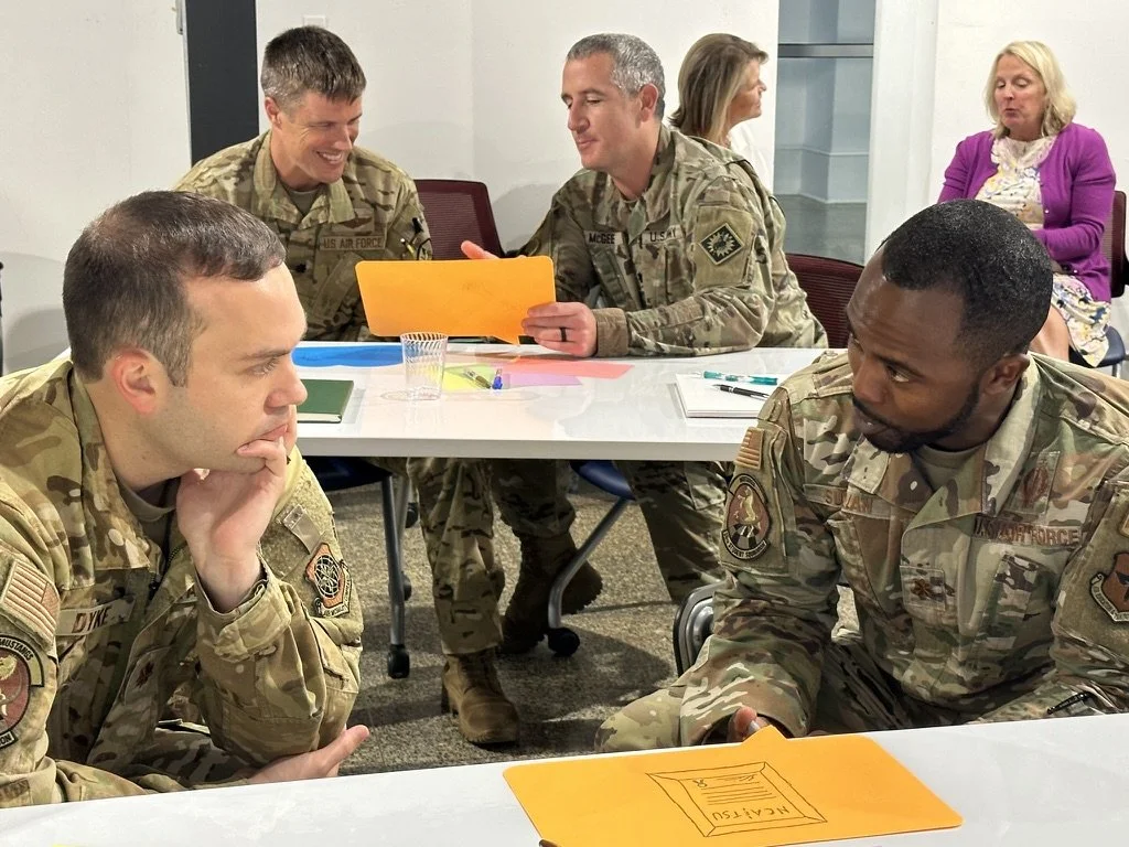 Four soldiers in uniform seated at tables, engaged in conversation, with two women sitting in the background. One soldier is resting his chin on his hand.