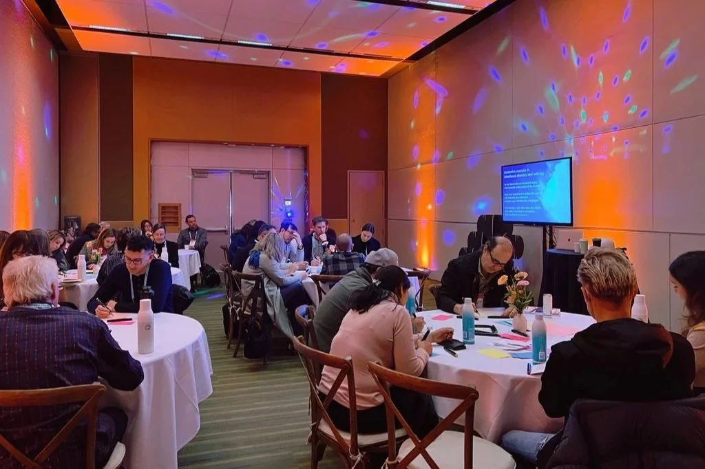 People sitting at round tables during a conference or workshop in a room with colorful lighting and a large screen displaying a presentation.