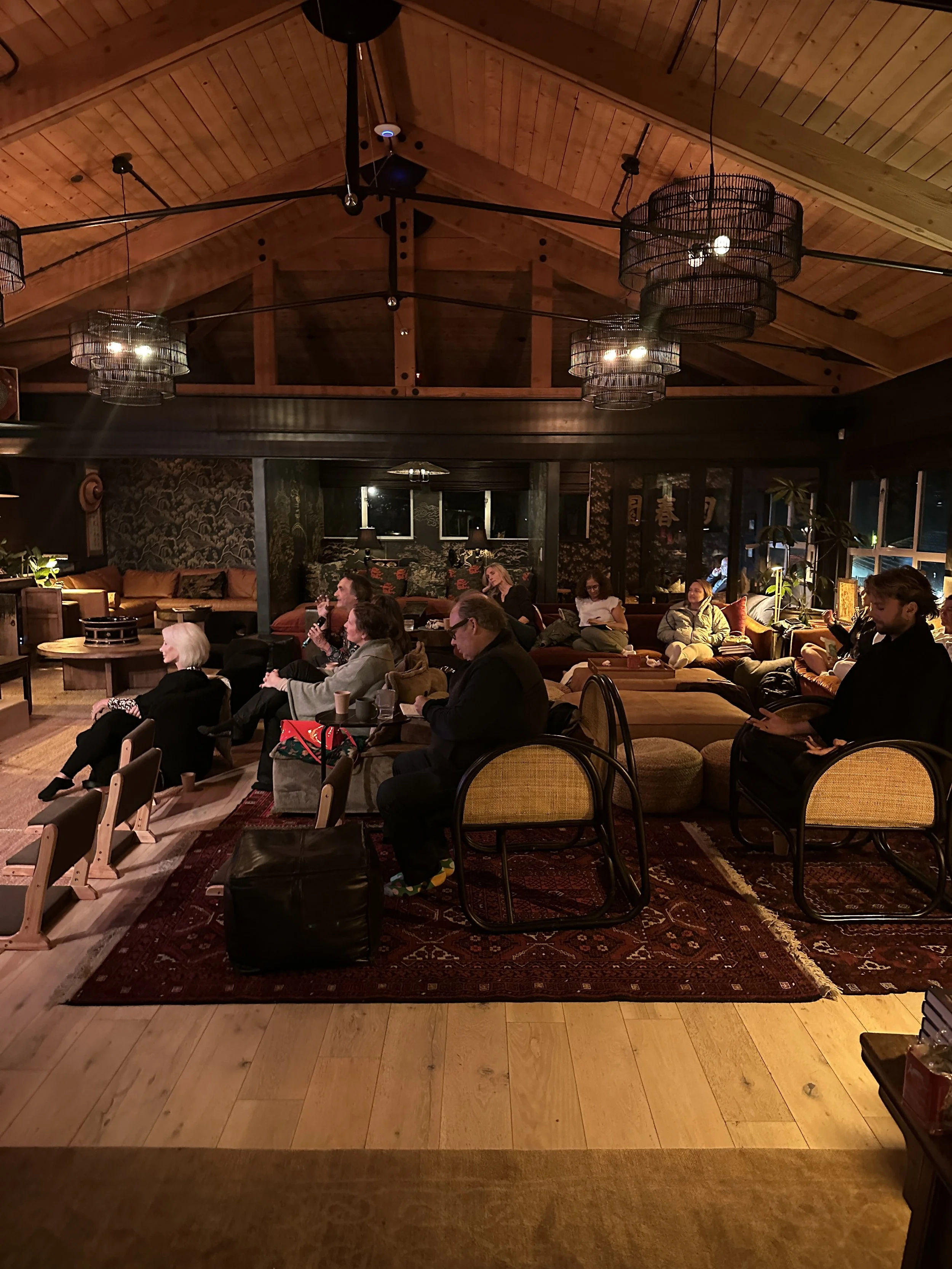 People seated in a cozy, warmly lit lounge or conference space with wooden ceiling and modern chandelier lighting, some engaged with their phones, others listening or drinking coffee.