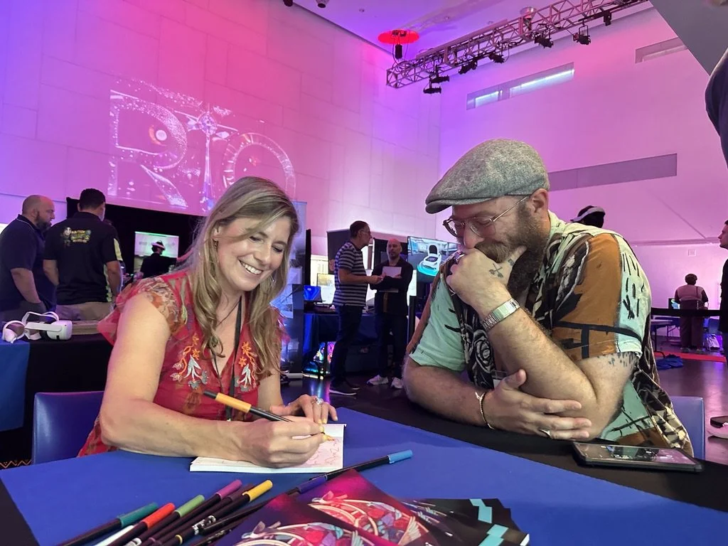 A woman with long blonde hair, wearing a red embroidered top, is smiling and signing an autograph at a table. A man with glasses, a gray flat cap, and a colorful t-shirt, with tattoos on his arm, is watching her sign with his chin resting on his hand