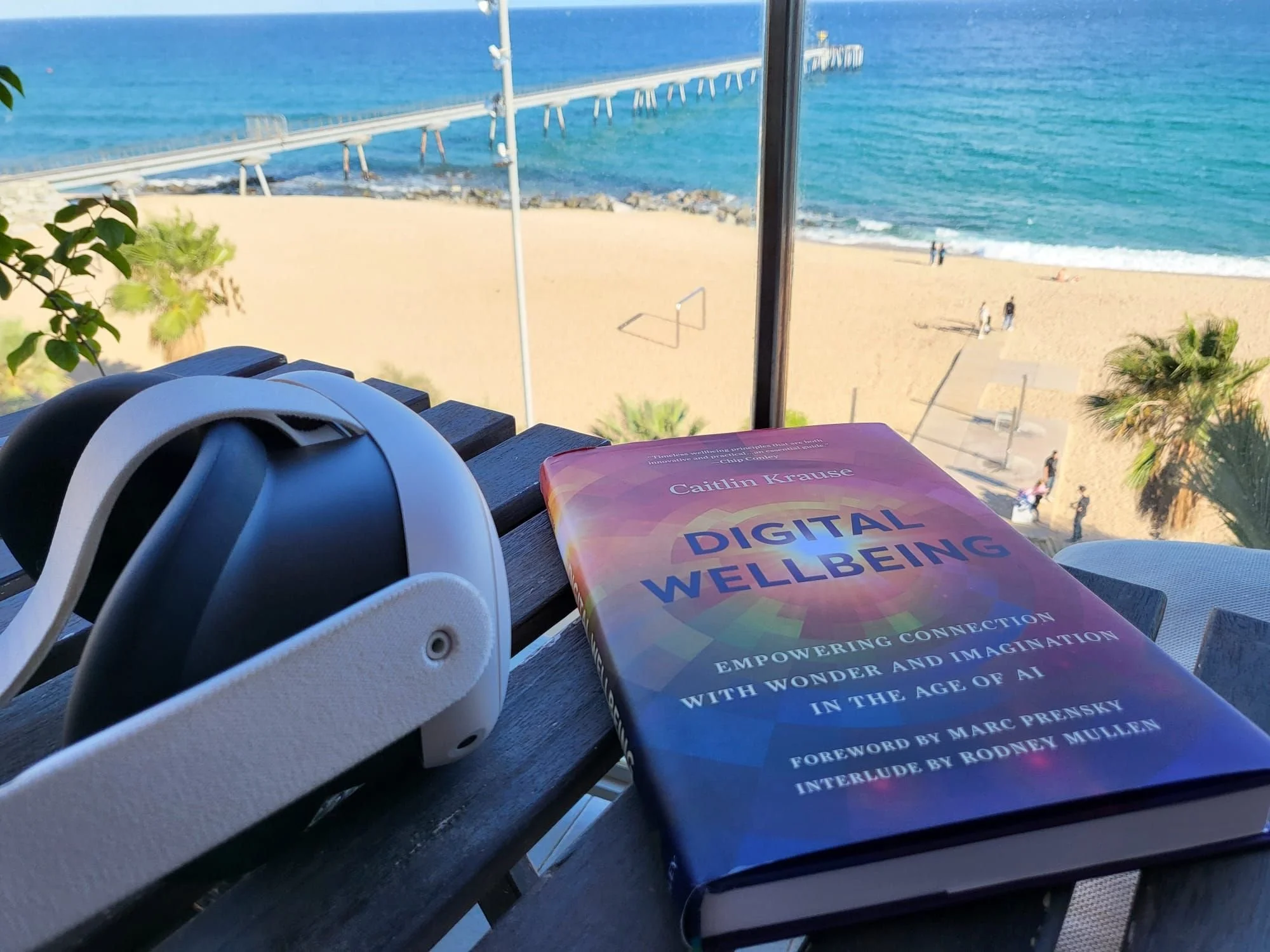 A book titled 'Digital Wellbeing' by Caitlin Krause, a pair of white headphones, and a view of a beach with sand, palm trees, and the ocean from a balcony or window.