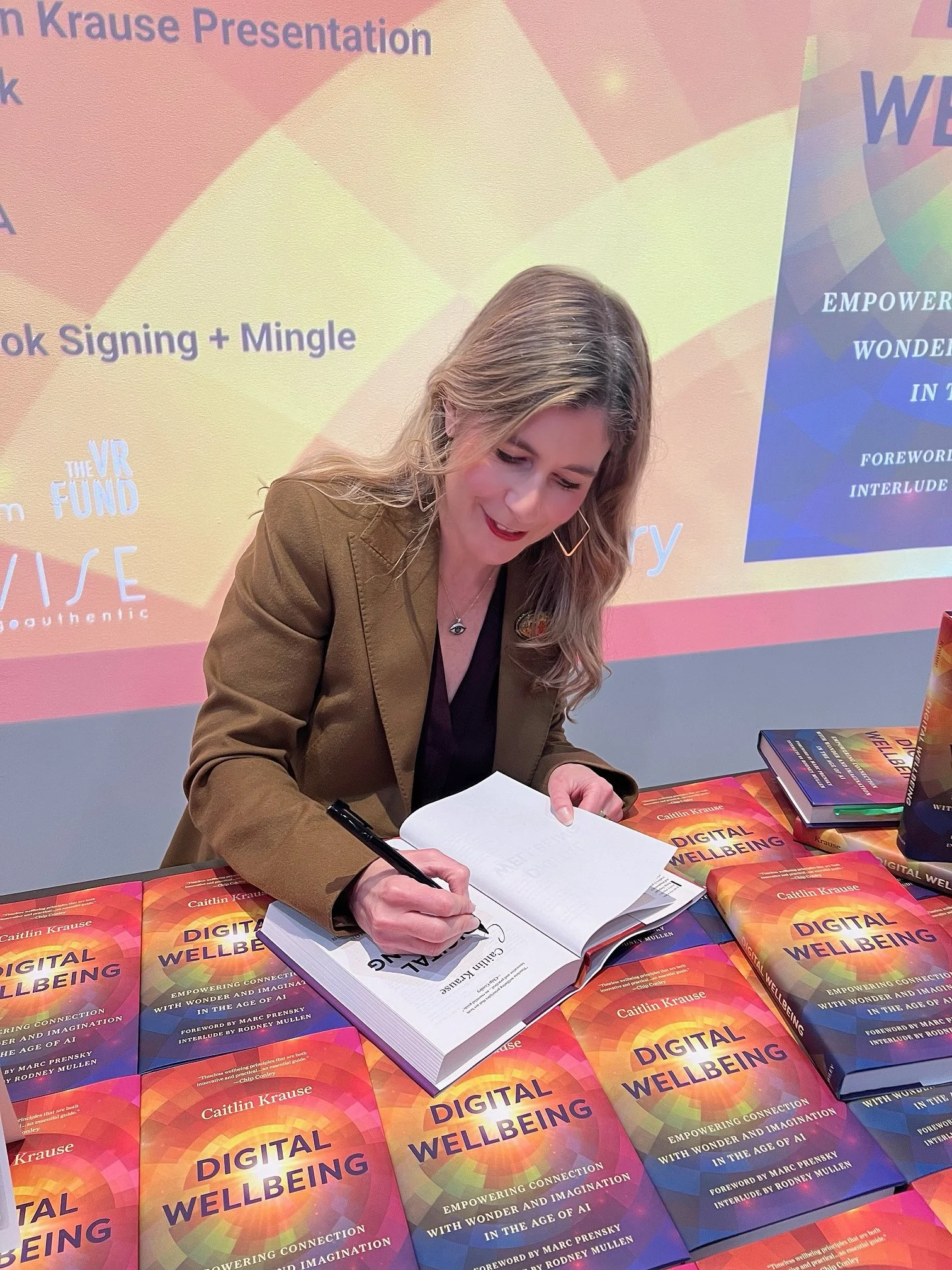 A woman (Caitlin Krause, author, keynote speaker) signing a book at a book signing event. Several copies of her book titled 'Digital Wellbeing' are displayed on the table in front of her. The background features a colorful presentation slide.