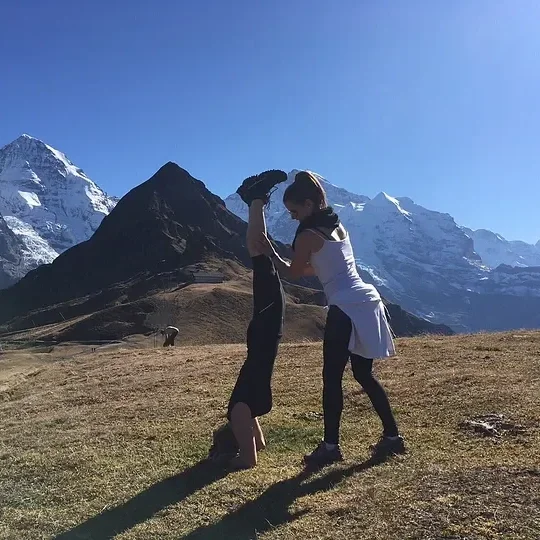 Two women (Caitlin Krause, author, keynote speaker) practicing acro yoga outdoors on a grassy plain with snow-capped mountains in the background.