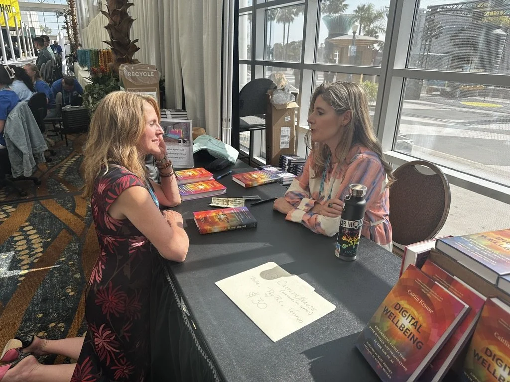 Two women sitting at a table during a book signing event, exchanging conversation. The table displays copies of a book titled 'Digital Wellbeing,' and a black water bottle with a colorful design. The setting is indoors with large windows showing an o