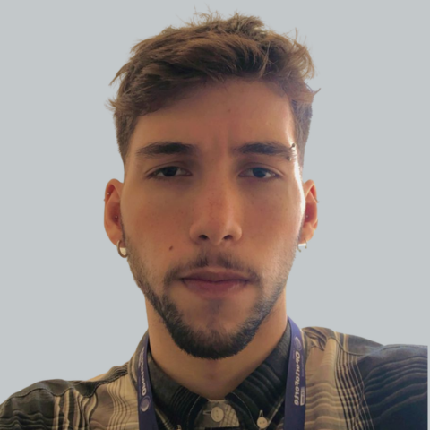 Jovem homem com cabelo castanho, barba e brinco, usando jaqueta com detalhes e credencial no pescoço