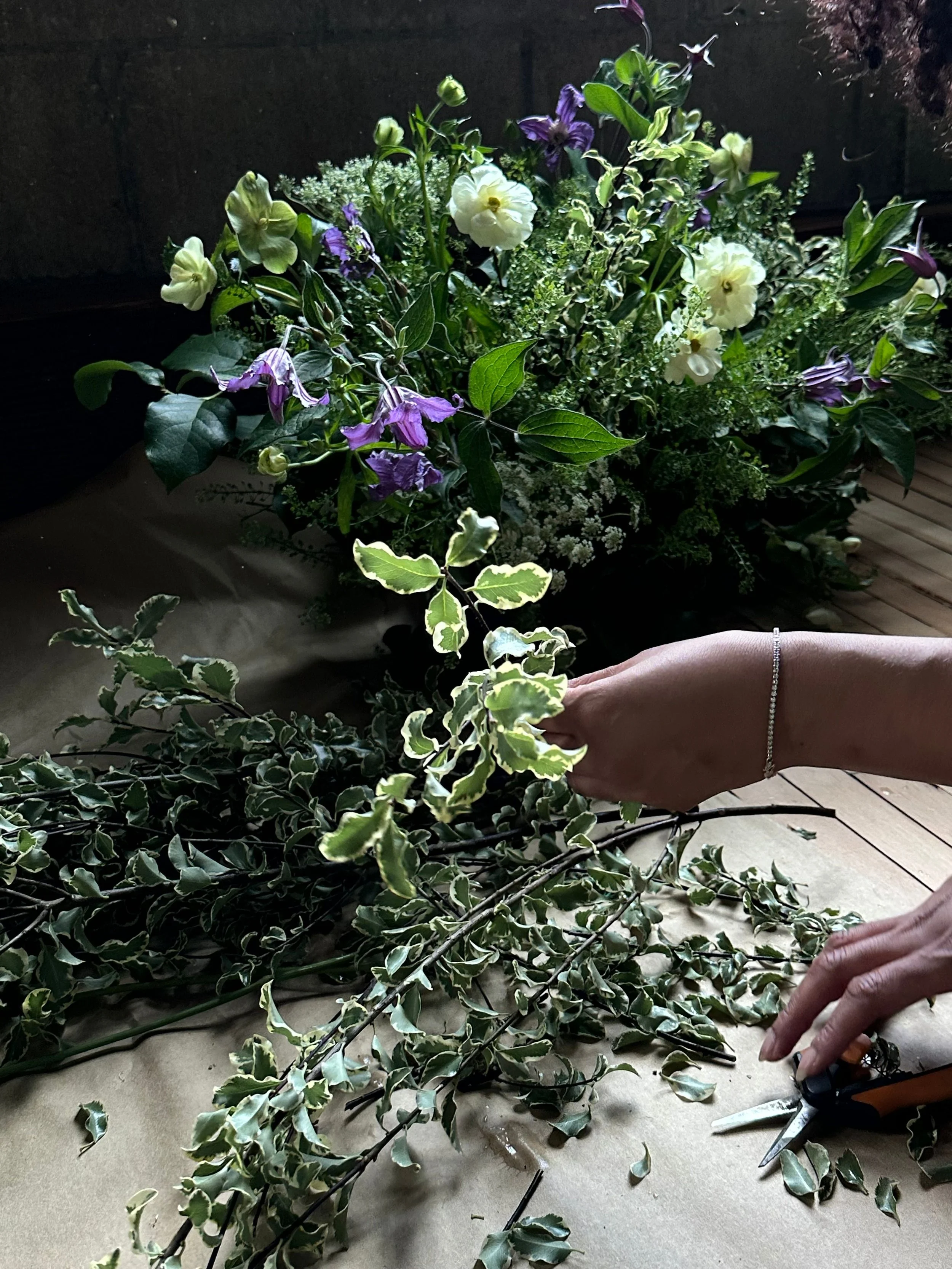 A person arranging a bouquet of assorted flowers and foliage on a table, with scissors nearby. The background shows a large arrangement of mixed flowers.