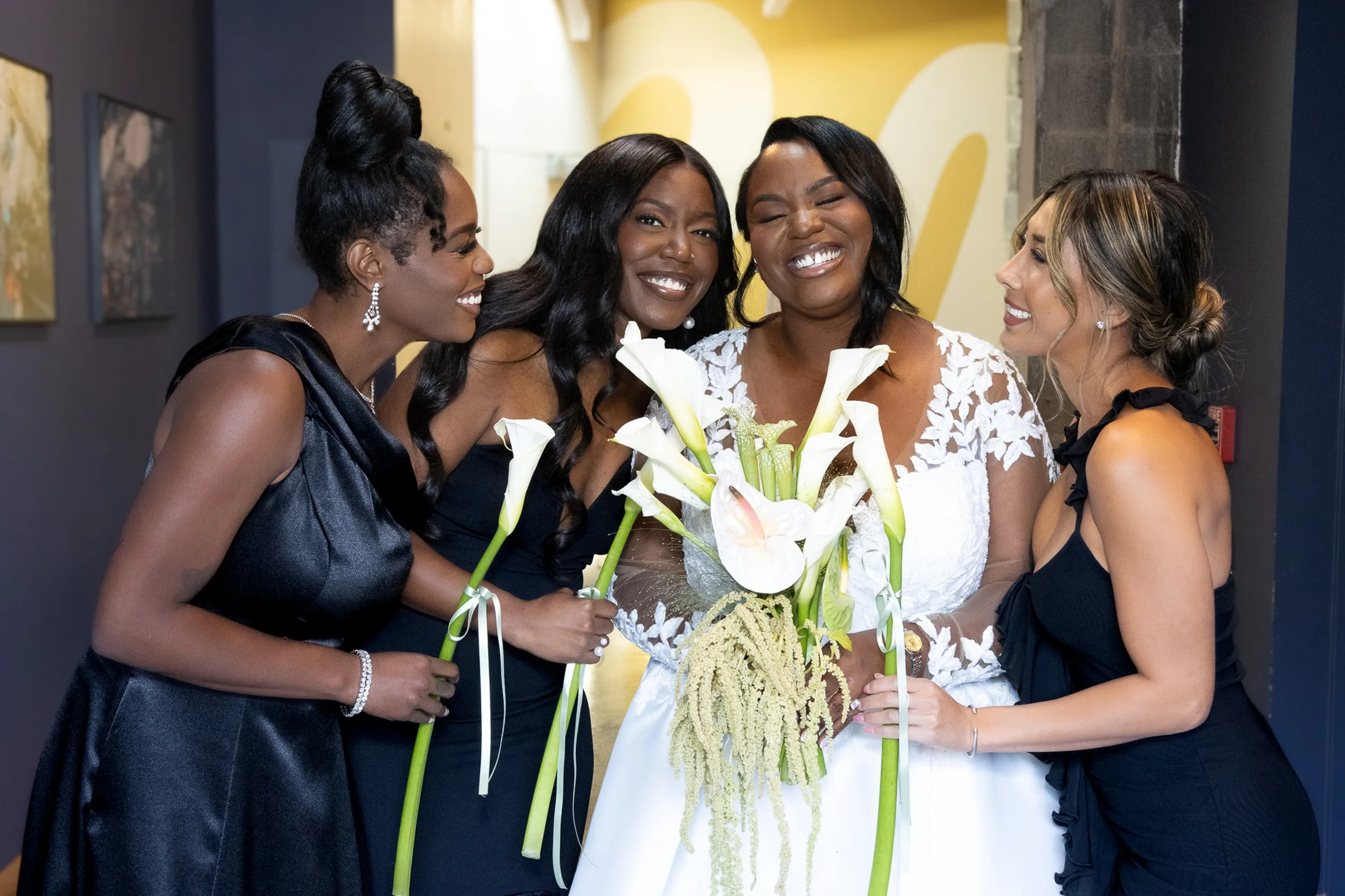 Four women dressed in elegant black and white dresses, smiling and holding bouquets of white calla lilies, standing together at a celebration or event.