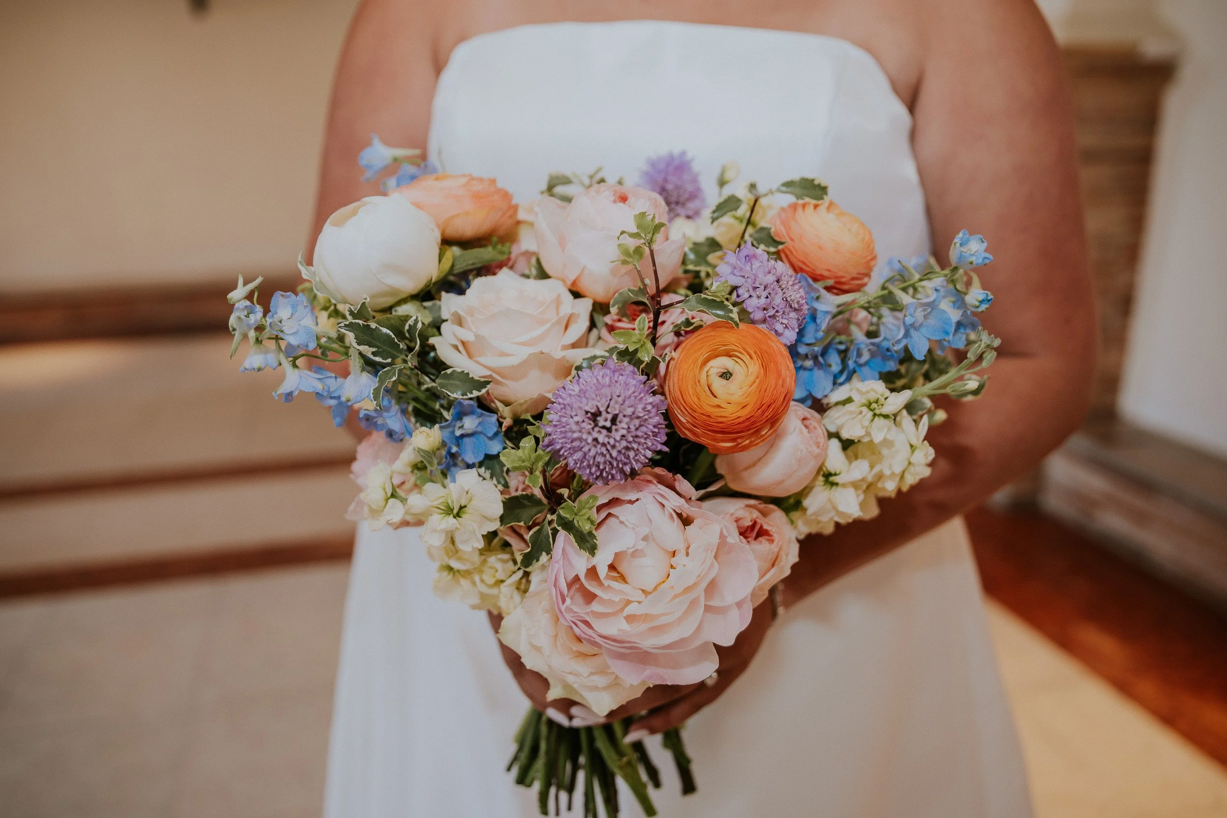 A person holding a large bouquet of multicolored flowers, including blush pink roses, orange ranunculus, blue delphiniums, and purple alliums, against a neutral background.