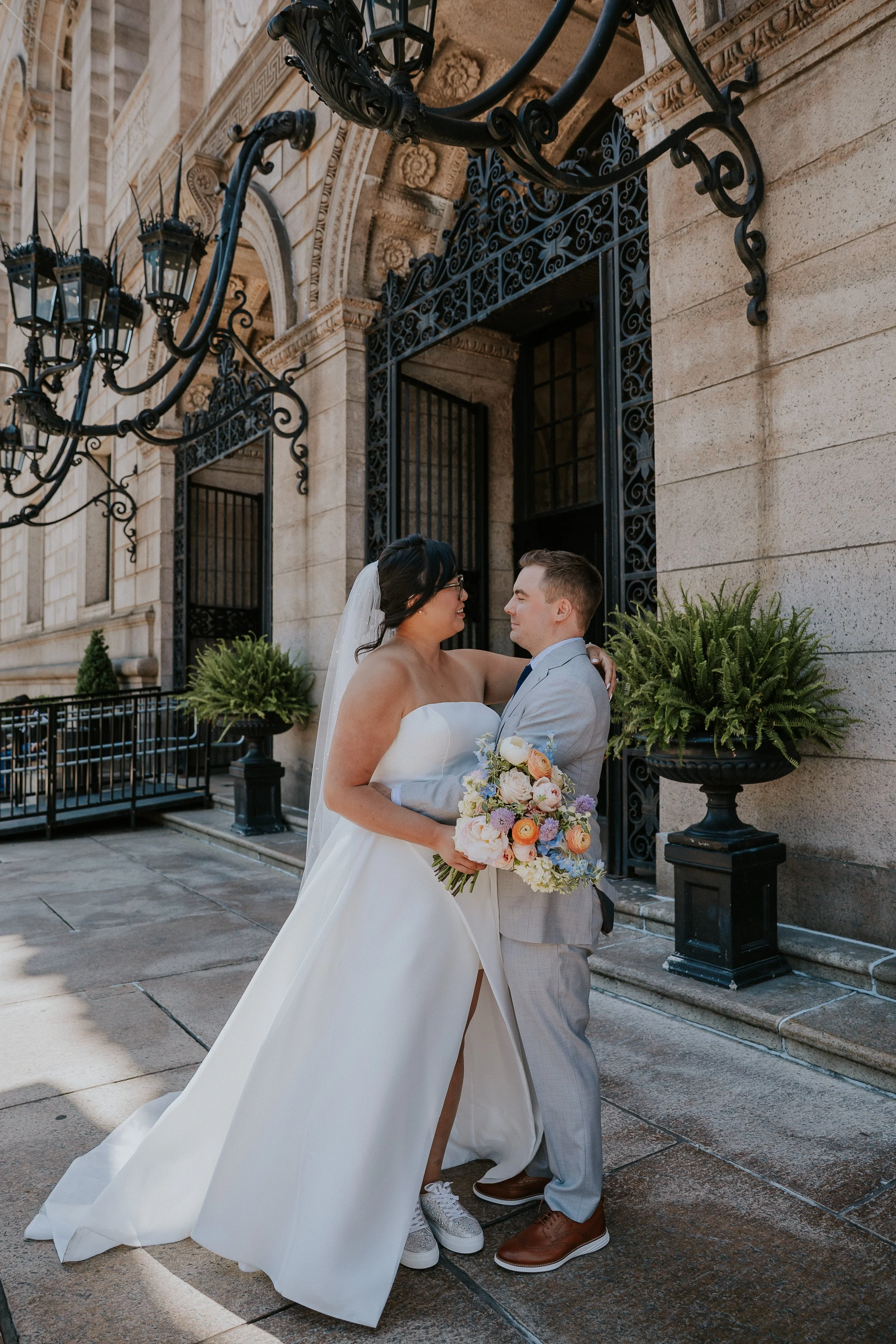 A bride and groom share a kiss outside a historic building, with the bride holding a bouquet of colorful flowers, and the groom wearing a light gray suit with brown shoes.