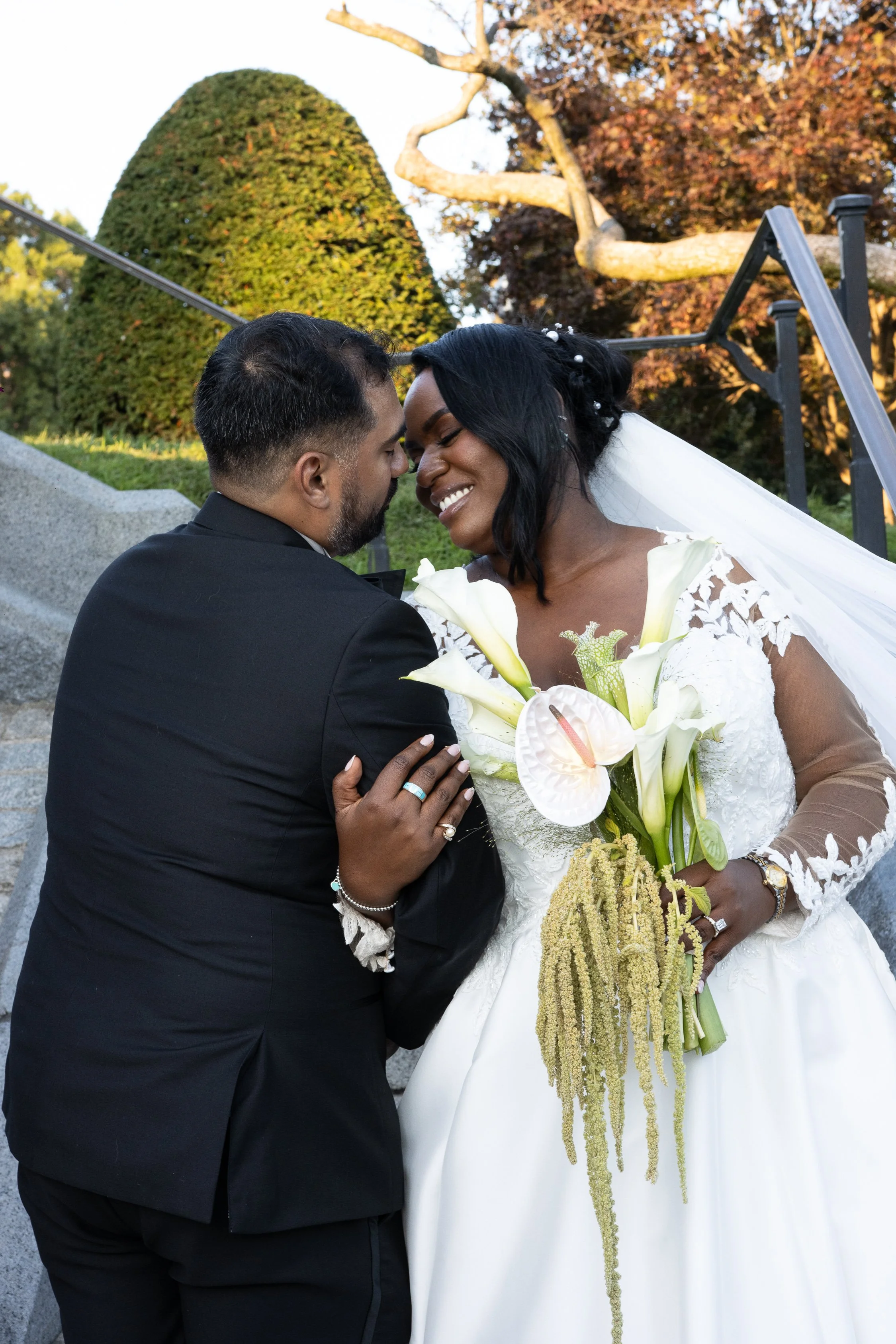 A bride and groom on their wedding day smiling and leaning close together outdoors with autumn trees and a hedge in the background.