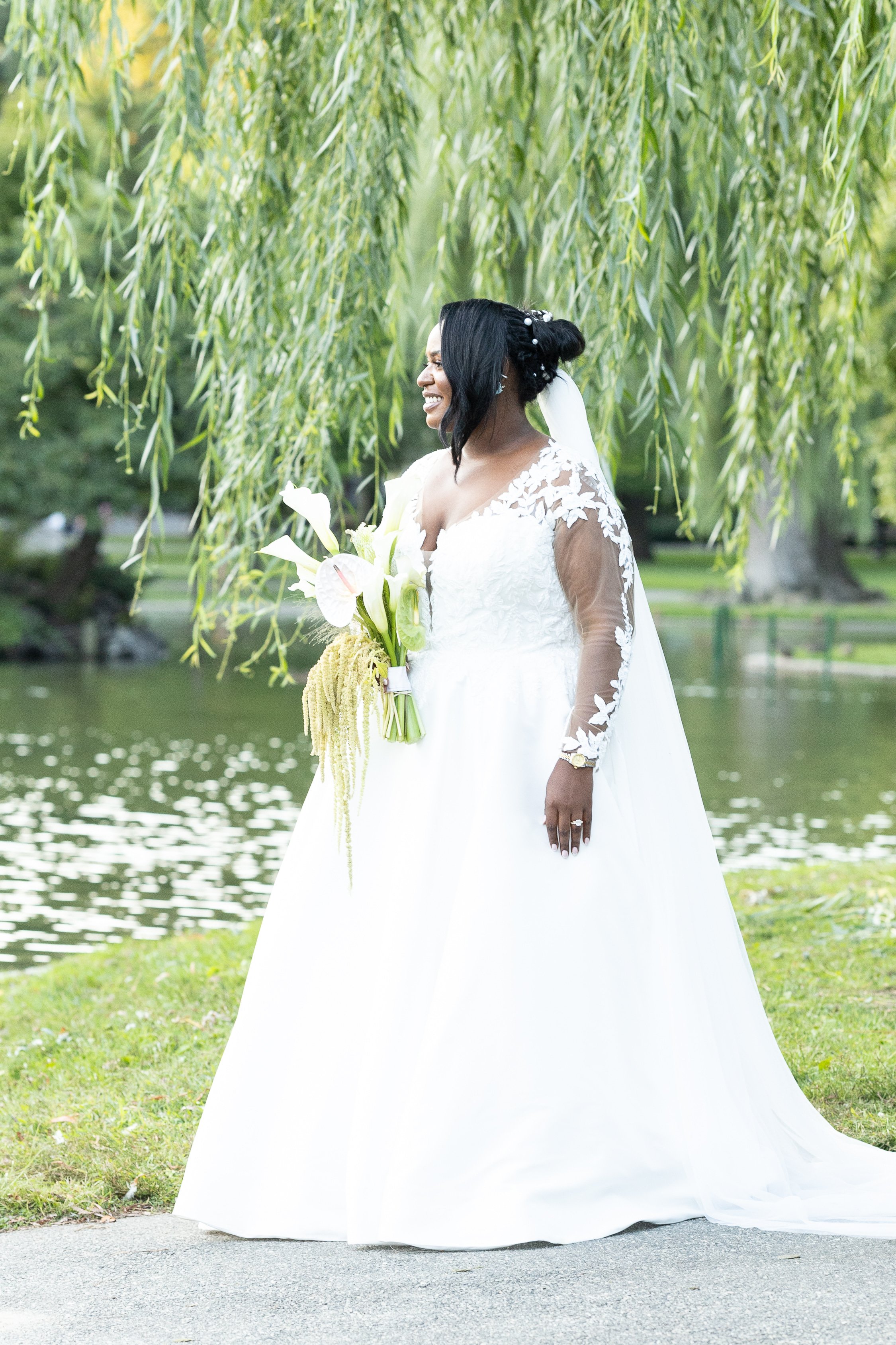 A bride in a white wedding gown holding a bouquet of lilies and greenery, smiling outdoors under a hanging tree near water.