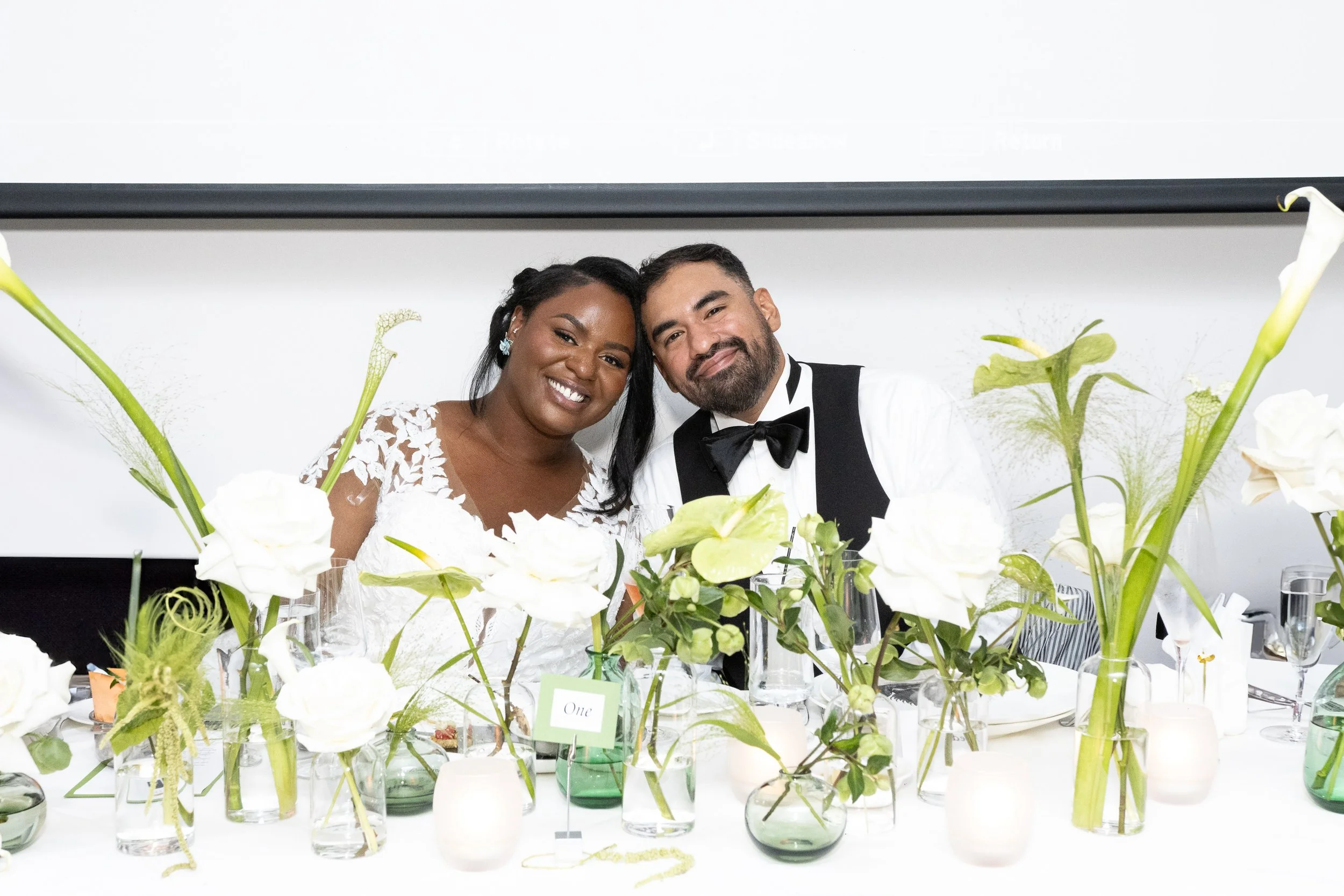 A smiling bride and groom at their wedding reception, sitting behind a table decorated with white flowers and green foliage.