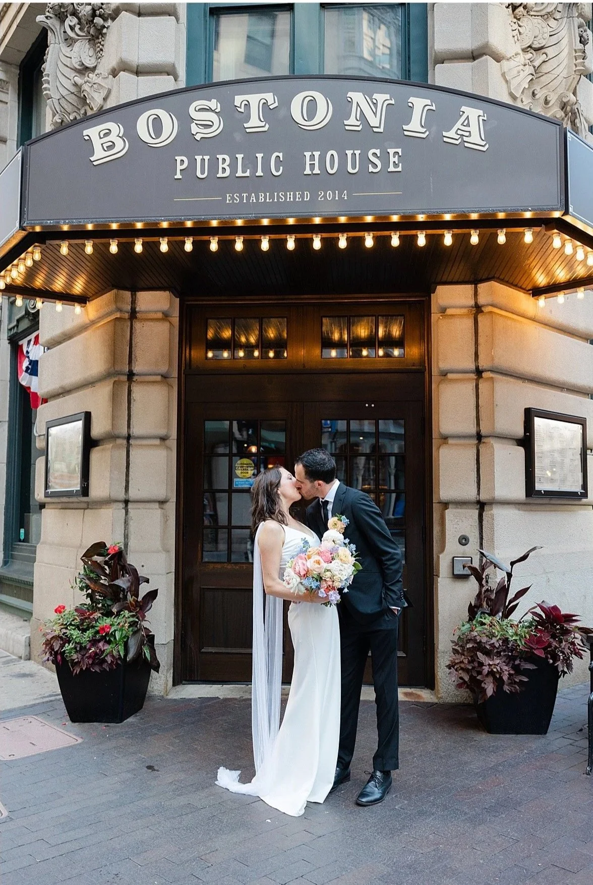 A newlywed couple sharing a kiss outside the Bostonia Public House, with the bride holding a bouquet of flowers, in front of the restaurant's entrance adorned with lighting and decorative planters.