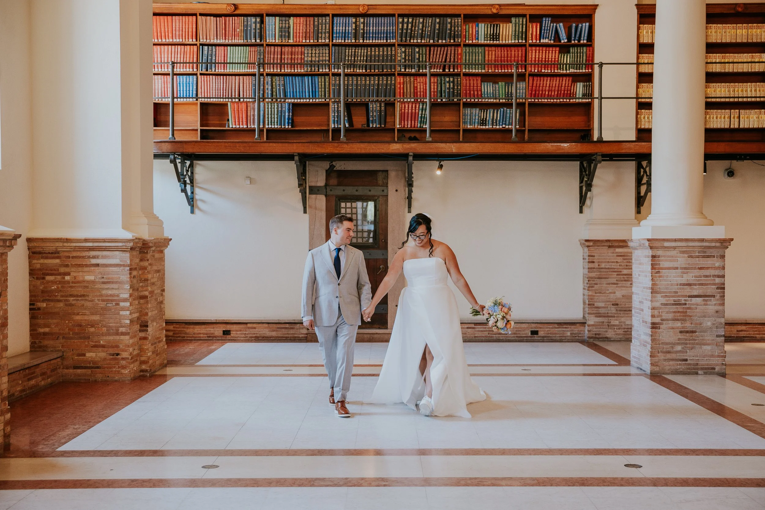 Bride and groom holding hands walking in a spacious room with bookshelves and brick columns.