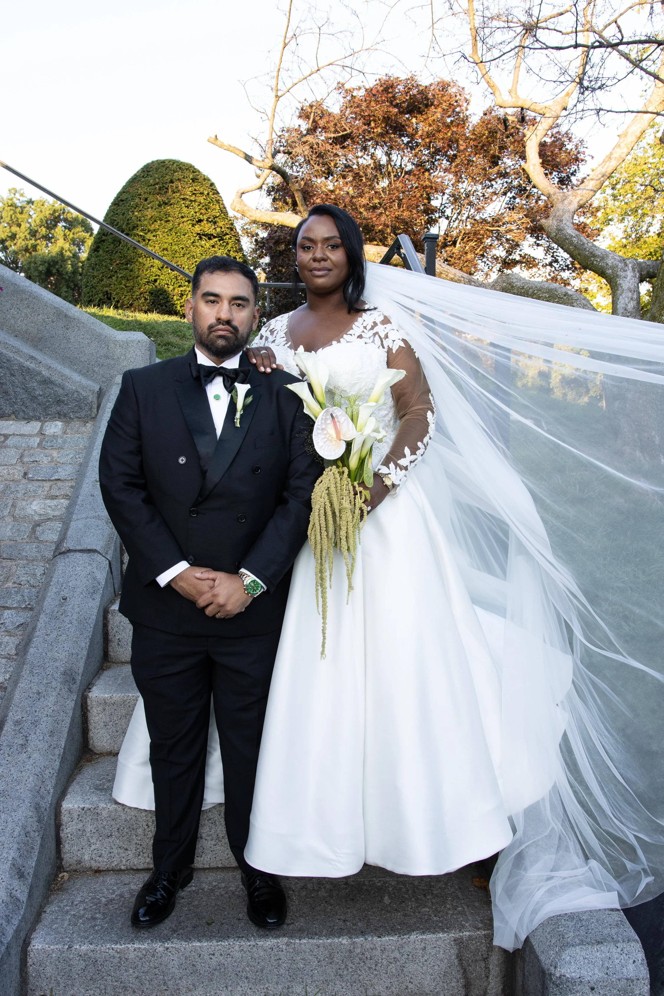 A bride and groom posing outdoors on stone steps during a wedding. The bride is wearing a white wedding gown with lace sleeves and holding a bouquet of white calla lilies. The groom is dressed in a black tuxedo with a bow tie. The background includes trees with autumn-colored leaves.