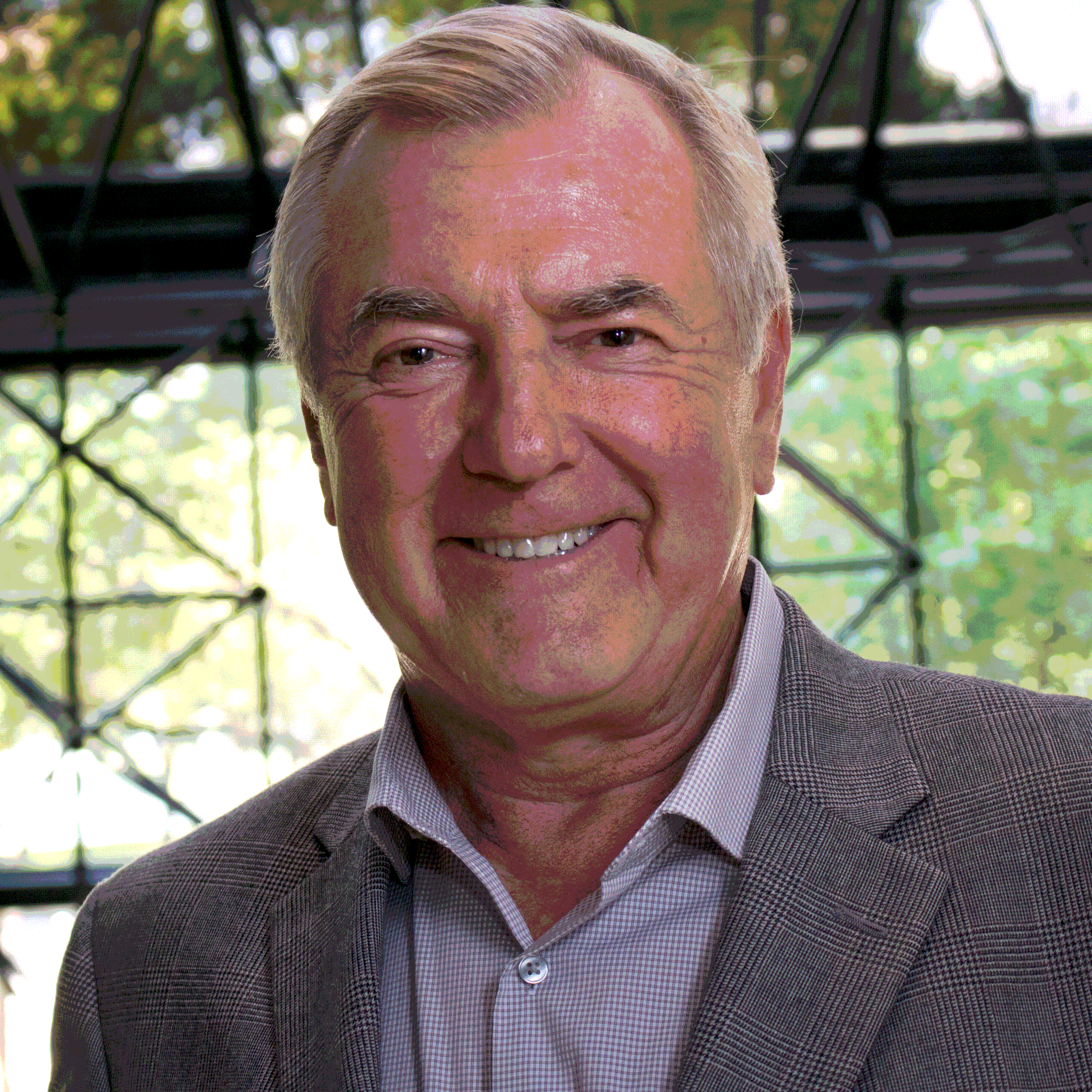 A middle-aged man with gray hair, smiling, wearing a gray checkered blazer and a white collared shirt. Background includes a large window with a metal grid and greenery outside.