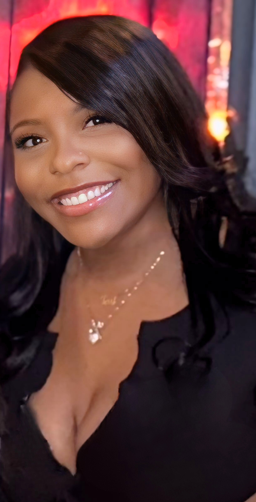 Close-up of a smiling woman with dark hair and wearing a black top, posing indoors with a warm, colorful background.