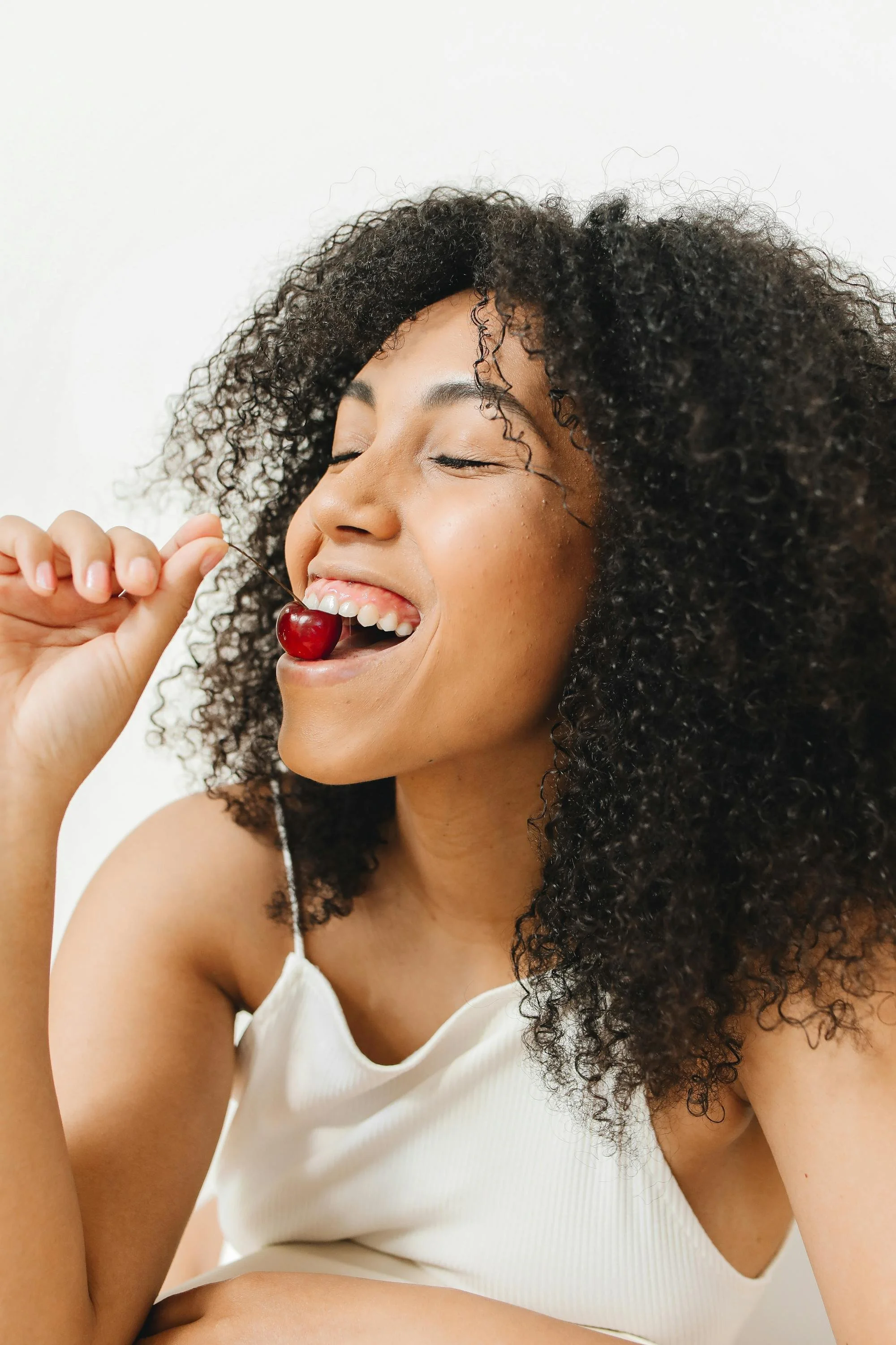 A woman with curly hair smiling with eyes closed, holding a cherry near her mouth.