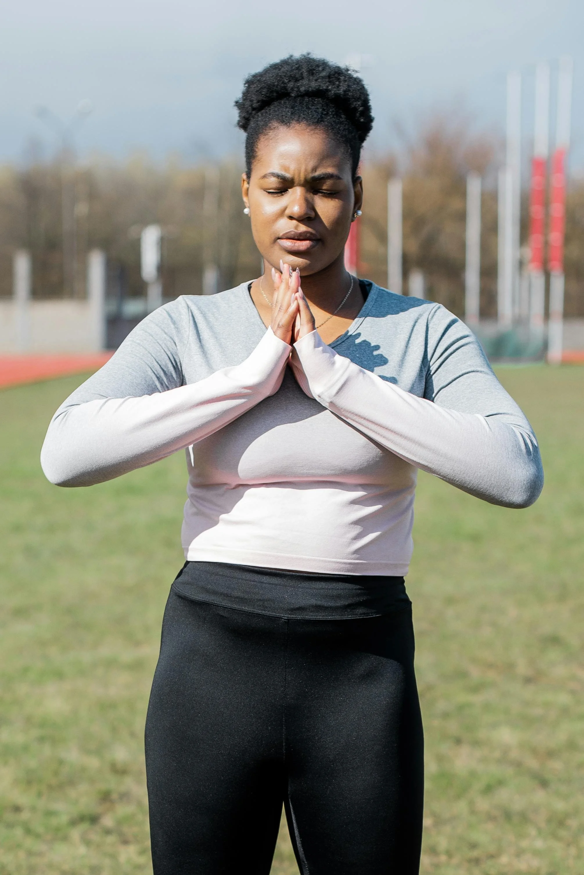 A young woman standing outdoors in athletic clothing, with her hands pressed together in a prayer position, eyes closed, appears to be praying or meditating on a sports field.