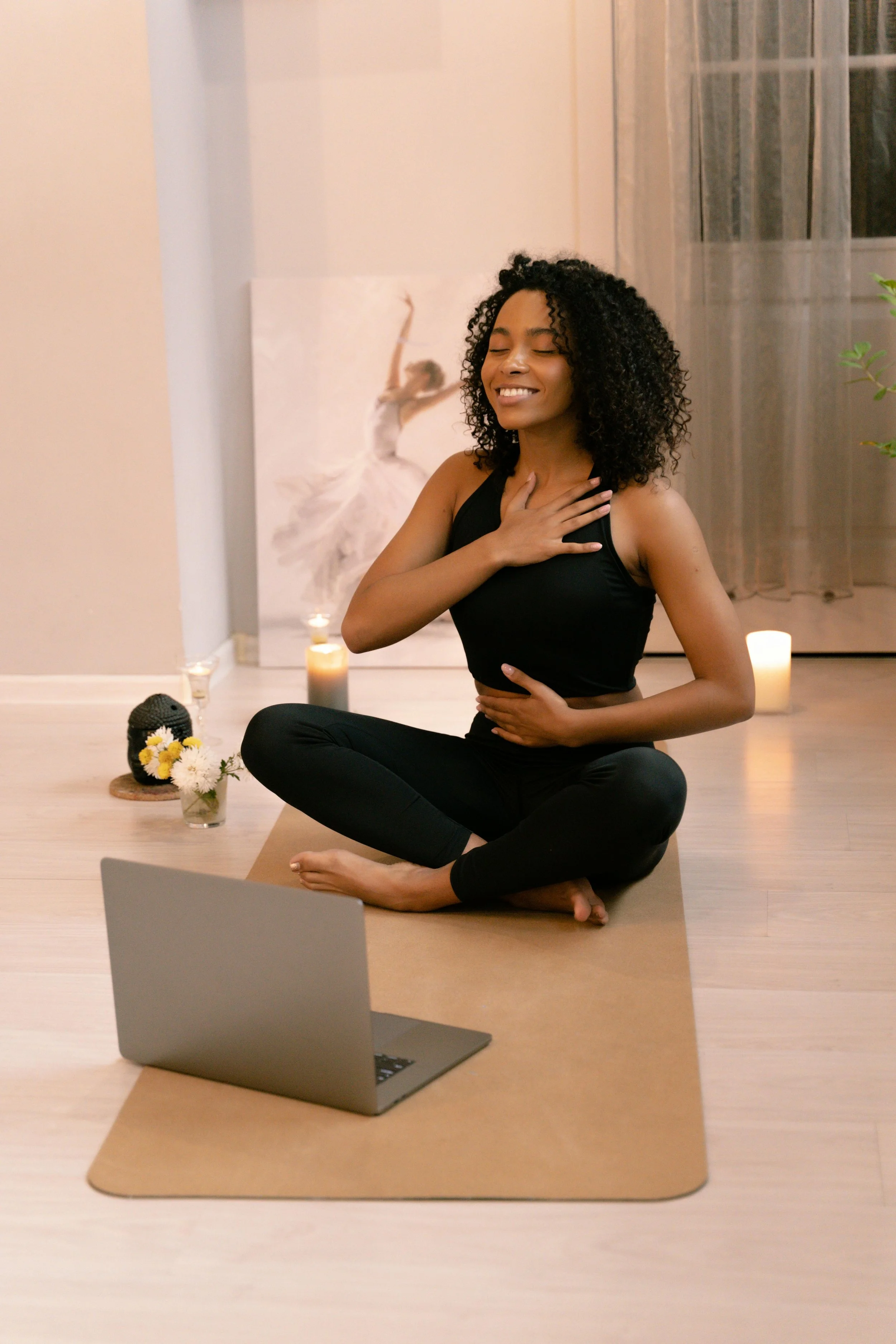 A woman practicing yoga in a cozy indoor setting, sitting cross-legged on a yoga mat with a laptop in front of her, surrounded by candles and decorative items.