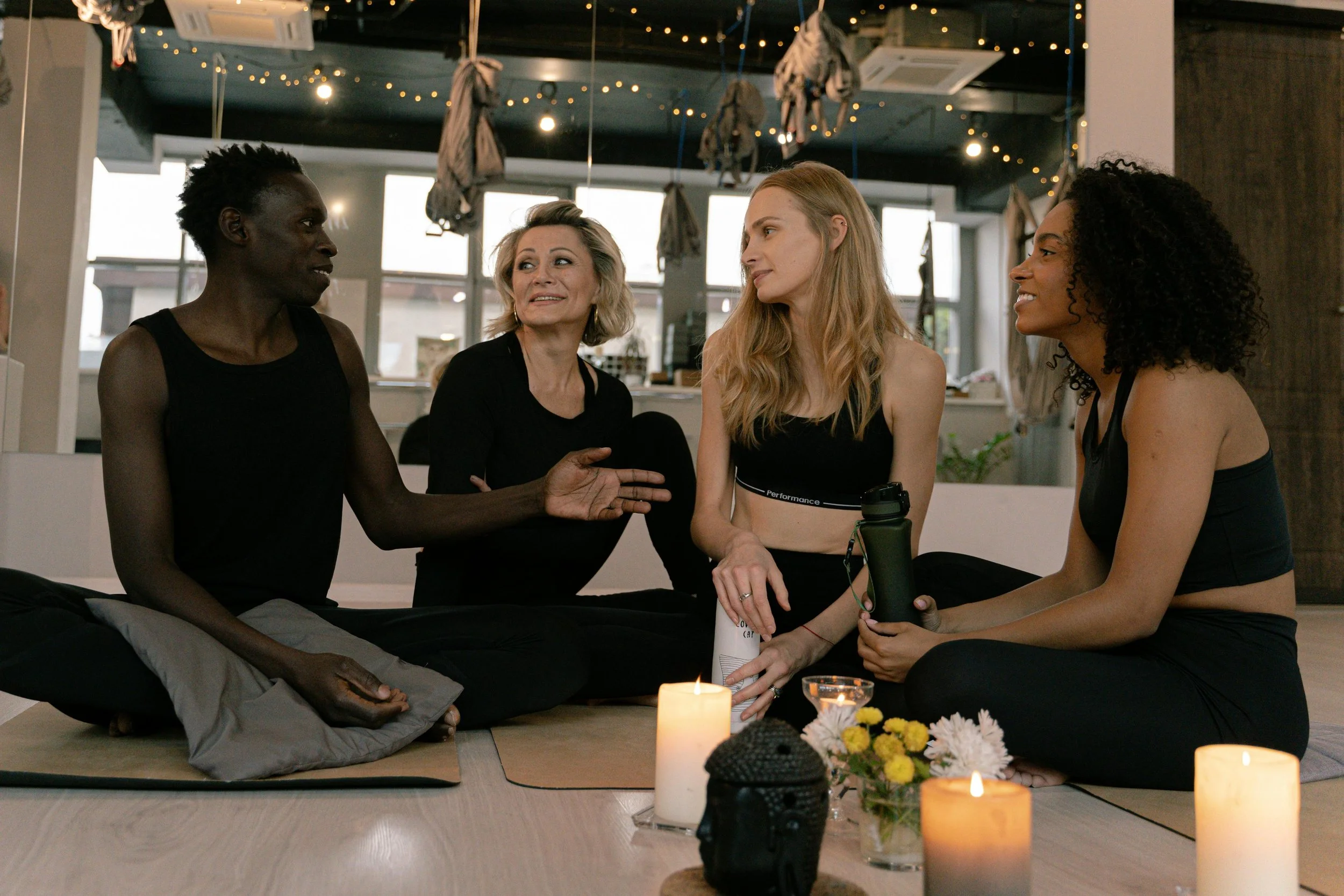 Four women sitting on the floor engaged in conversation, with candles and flowers in front of them, inside a cozy, well-lit room