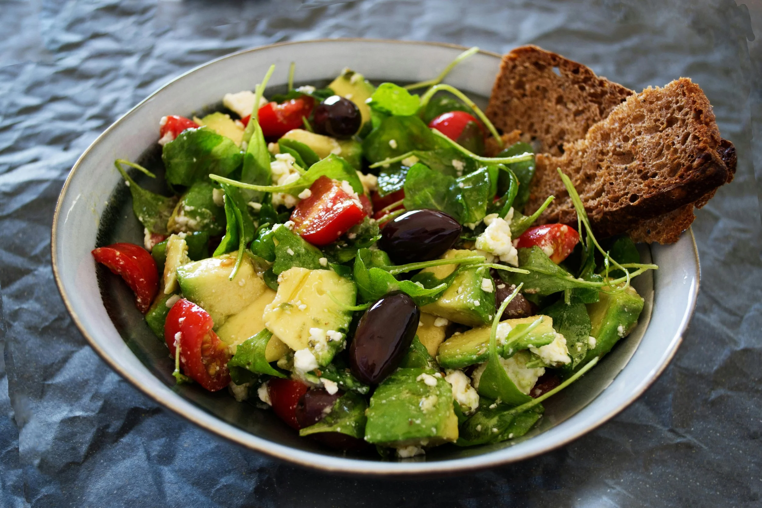 Bowl of salad with cherry tomatoes, avocado, green leafy vegetables, crumbled cheese, and black olives, served with two slices of whole grain bread.