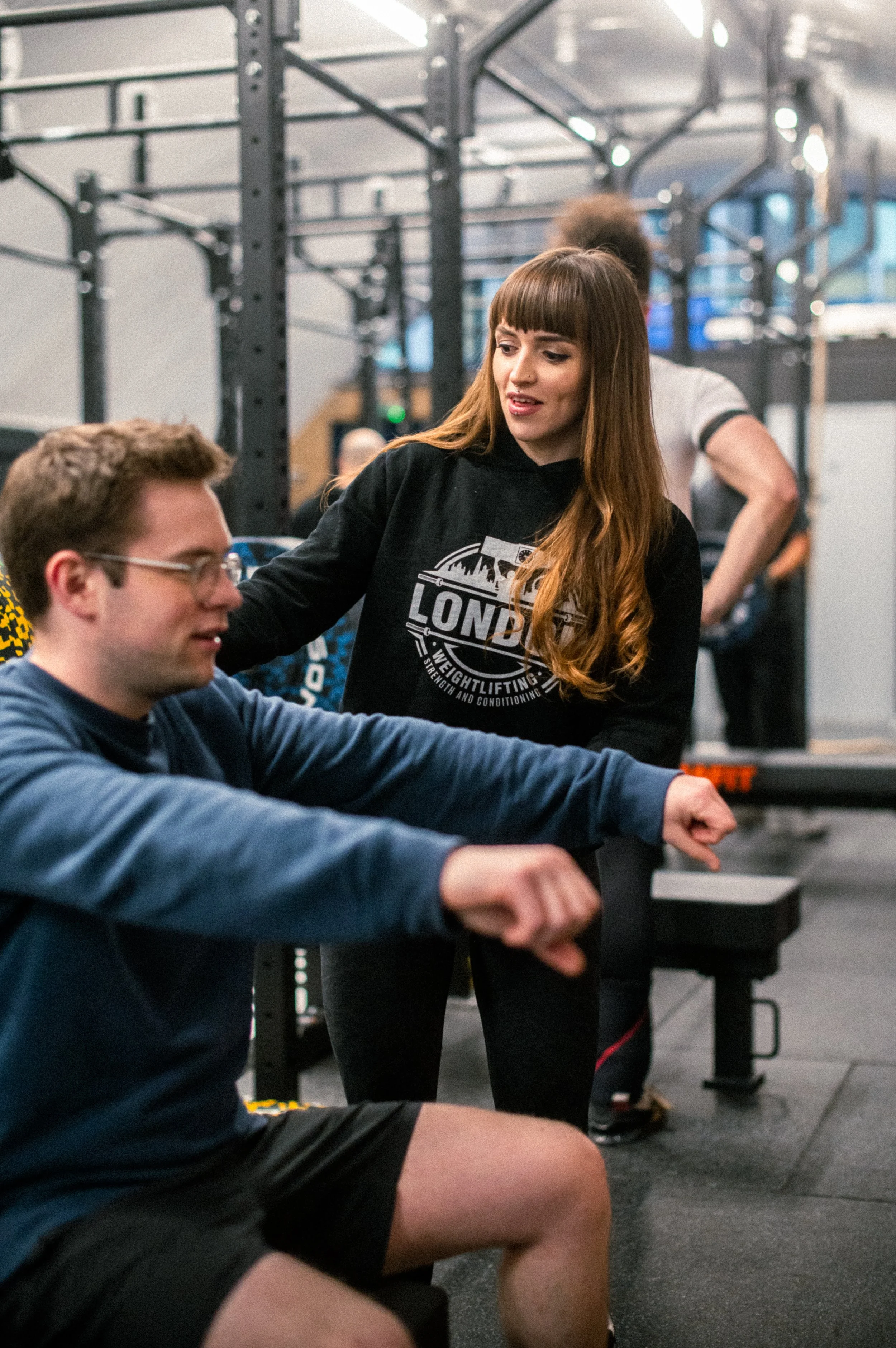 A woman with long red hair assists a seated man in a gym, both are arm-wrestling, with gym equipment in the background.