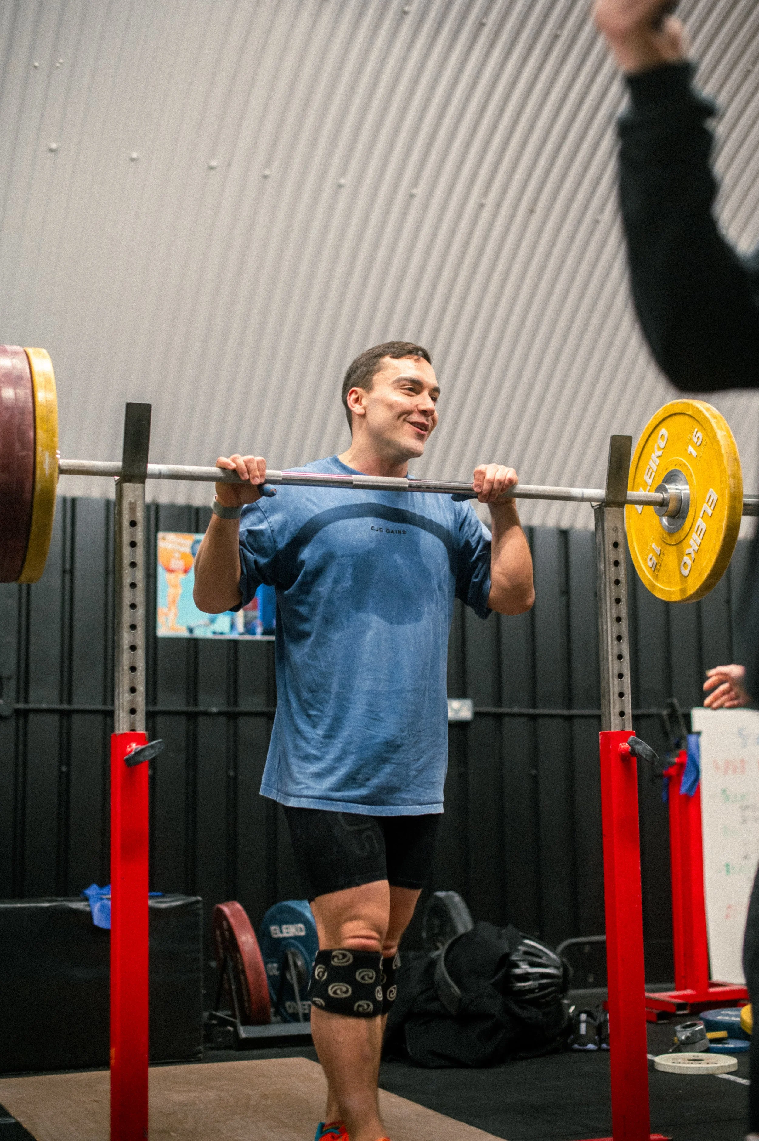 A man lifting a barbell with weights in a gym, smiling, with a dark background and workout equipment around him.