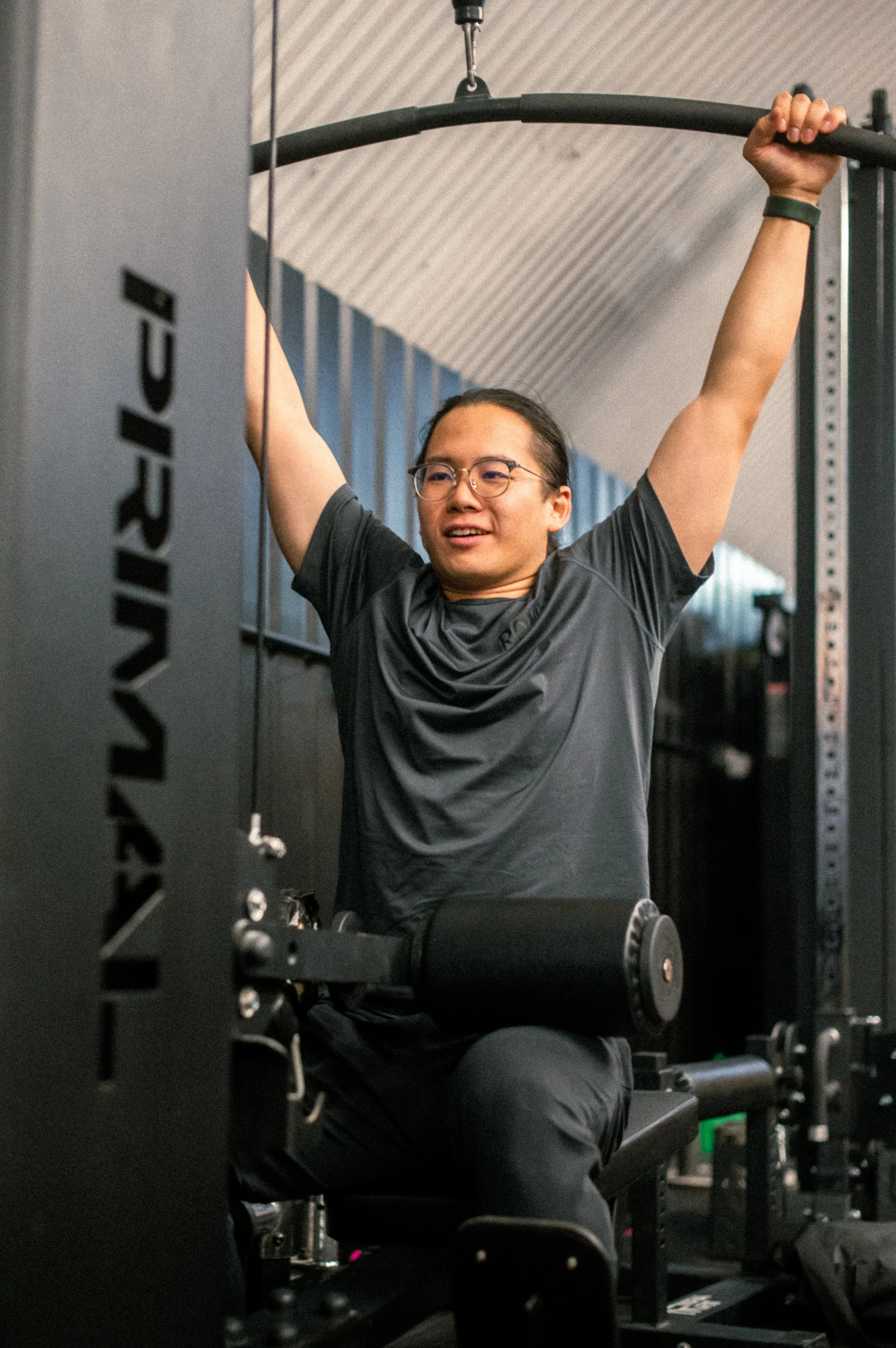 Man exercising on a fitness machine at gym, wearing glasses and a gray t-shirt, with arms raised holding a horizontal bar.