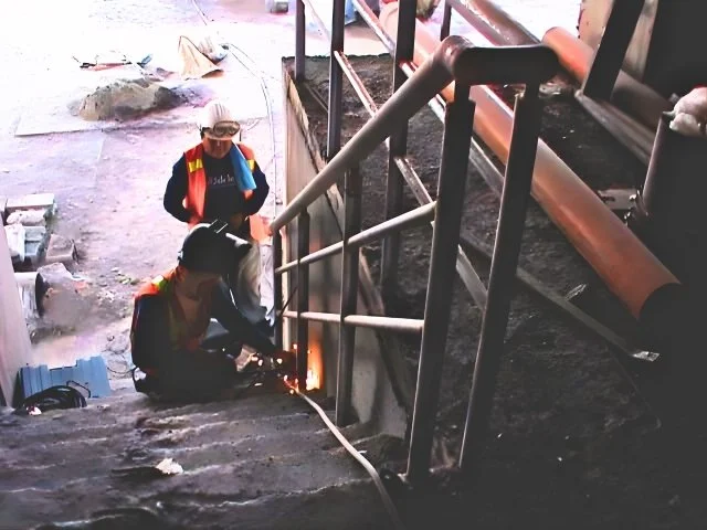 Two workers in safety gear welding a metal railing on a staircase at an industrial site.