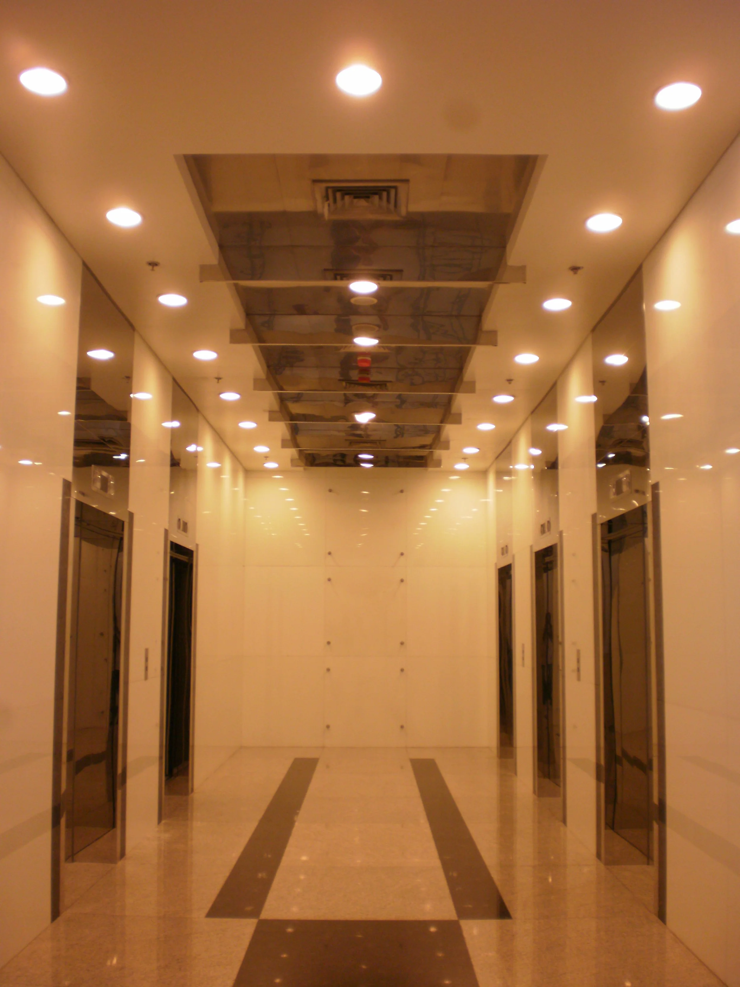 Empty elevator lobby with four elevators on each side, polished floors, beige walls, and overhead lighting.