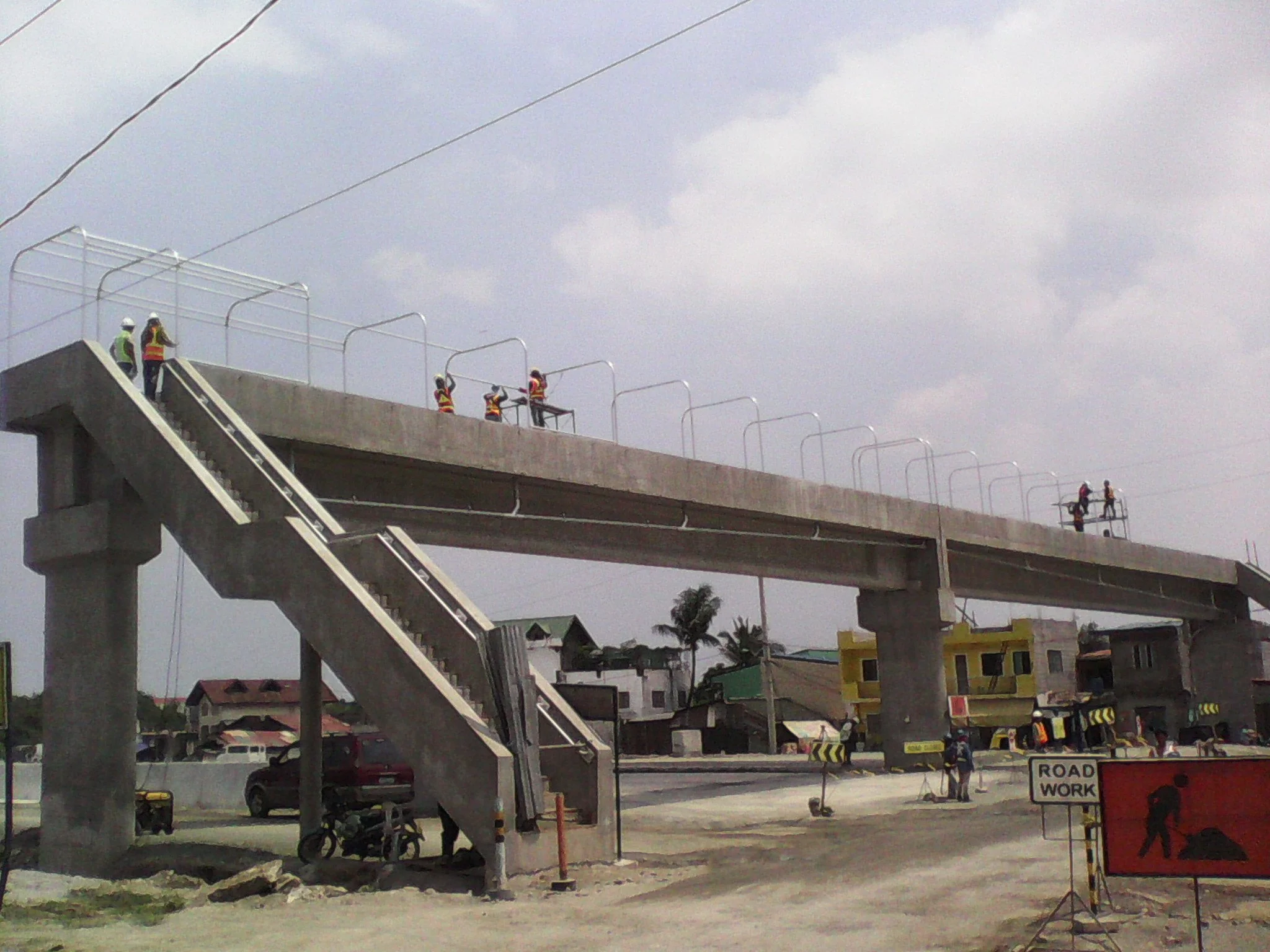 Construction workers working on a new overpass bridge, some stand on scaffolding and others walk on the bridge, with roadwork signs and equipment nearby.
