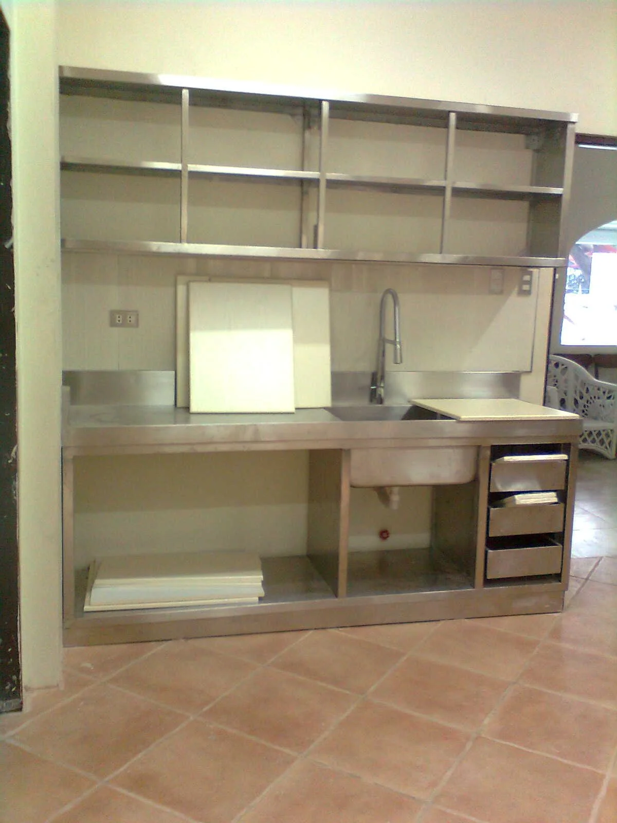 Empty stainless steel commercial kitchen sink station with open shelves above, a faucet, and a small built-in storage area below on a tiled floor.