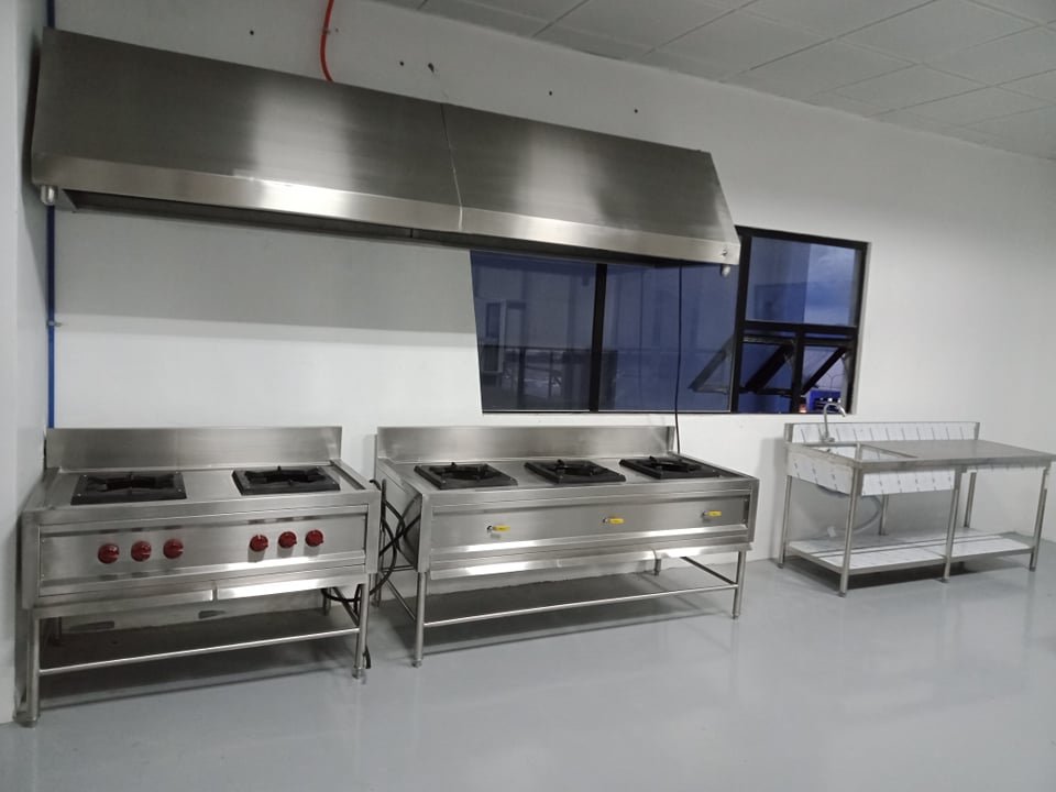 Commercial kitchen with two gas stoves, a large stainless steel hood, a stainless steel prep table, and a window with a black frame.