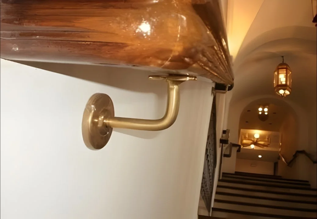 Close-up of a brass handrail support attached to a wooden handrail on a staircase in a warmly lit interior.