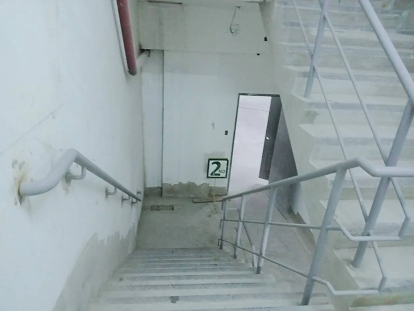 A stairwell in a building with beige walls, a metal handrail on the left, and a metal railing on the right. The stairs are concrete, and there is a door at the bottom of the stairs with a sign indicating "2nd" floor. The image appears to be taken from the top of the stairs looking down.