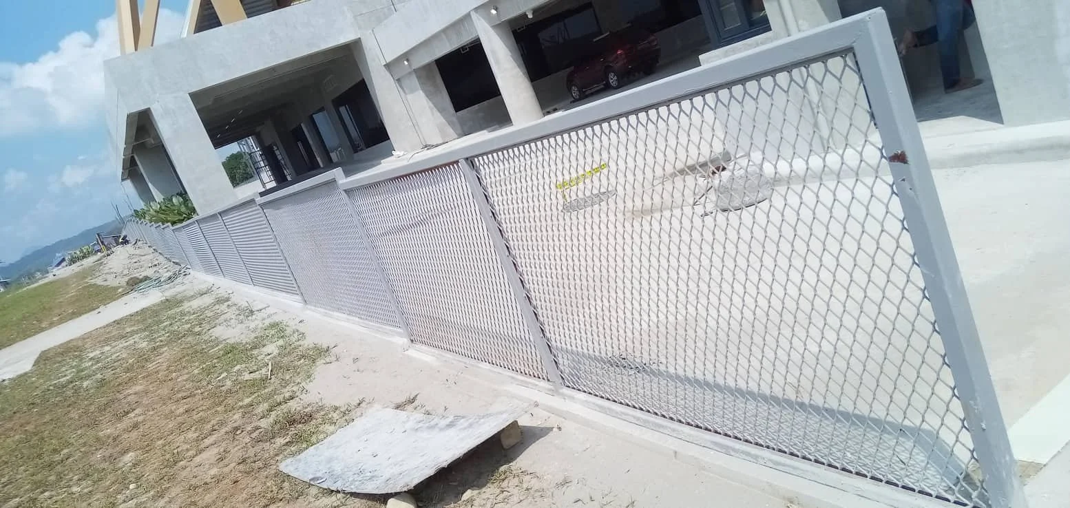 White metal fence in front of a concrete building under construction, with a sidewalk and grassy area beside it, and parked cars visible through the building's open garage.