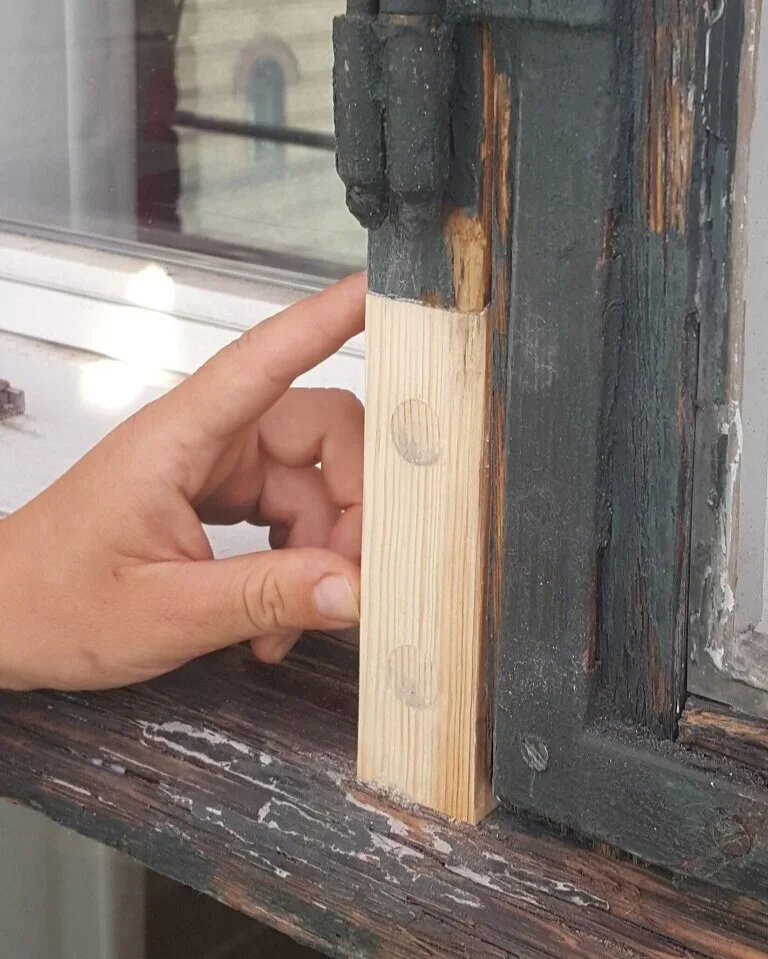 En hånd holder et stykke træ mod en drejestang i en vinduesramme, sandsynligvis til reparation eller montering.