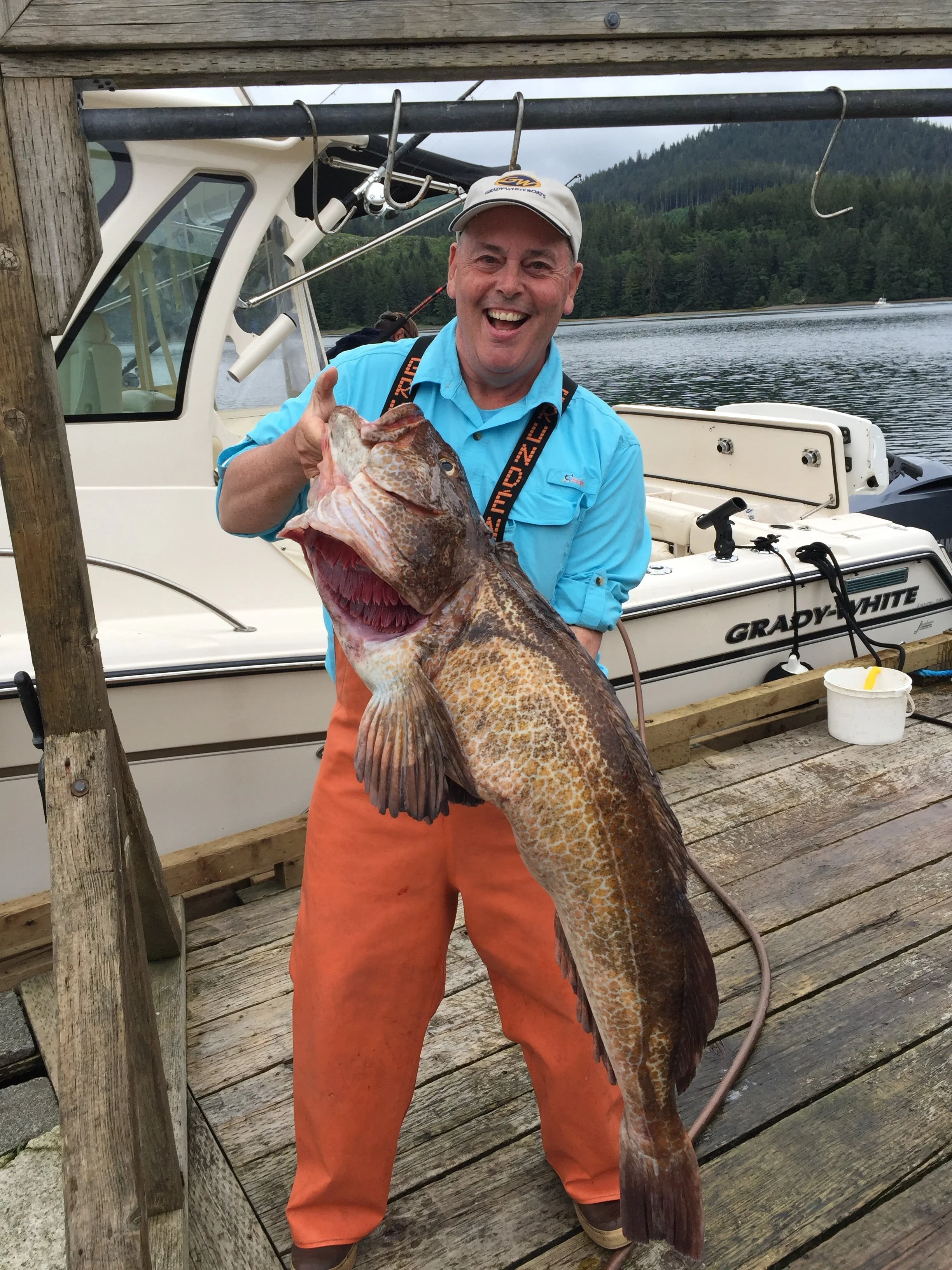 A man smiling and holding a large fish on a dock near a boat, with a lake and forested hills in the background.