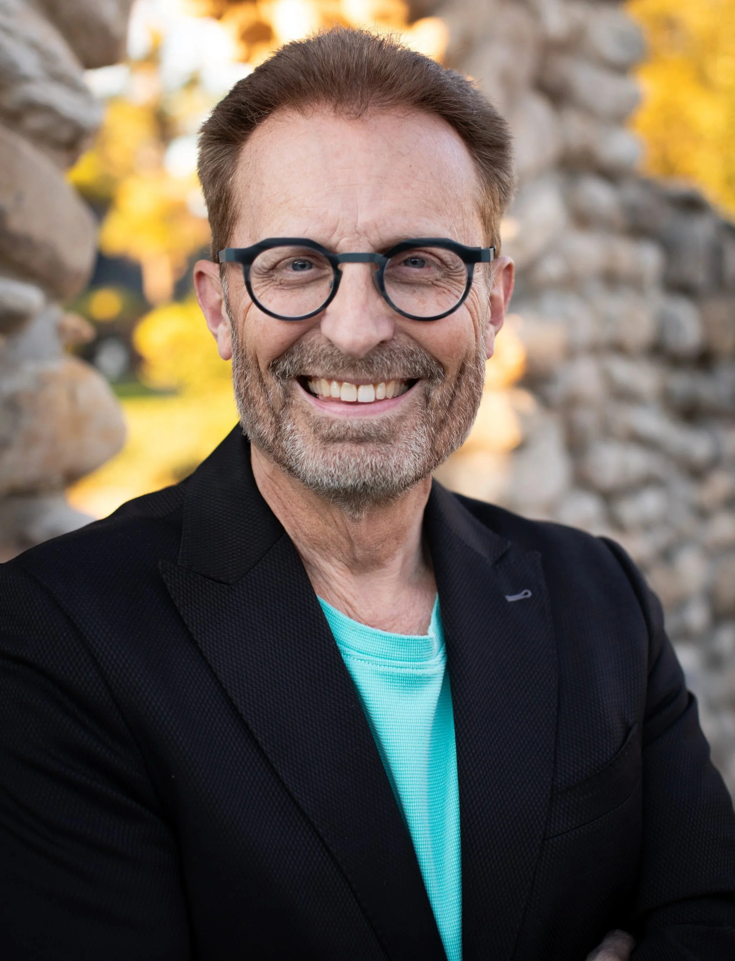 Close-up of a smiling middle-aged man wearing glasses, a black jacket, and a turquoise shirt, standing outdoors between stone walls with autumn-colored trees in the background.
