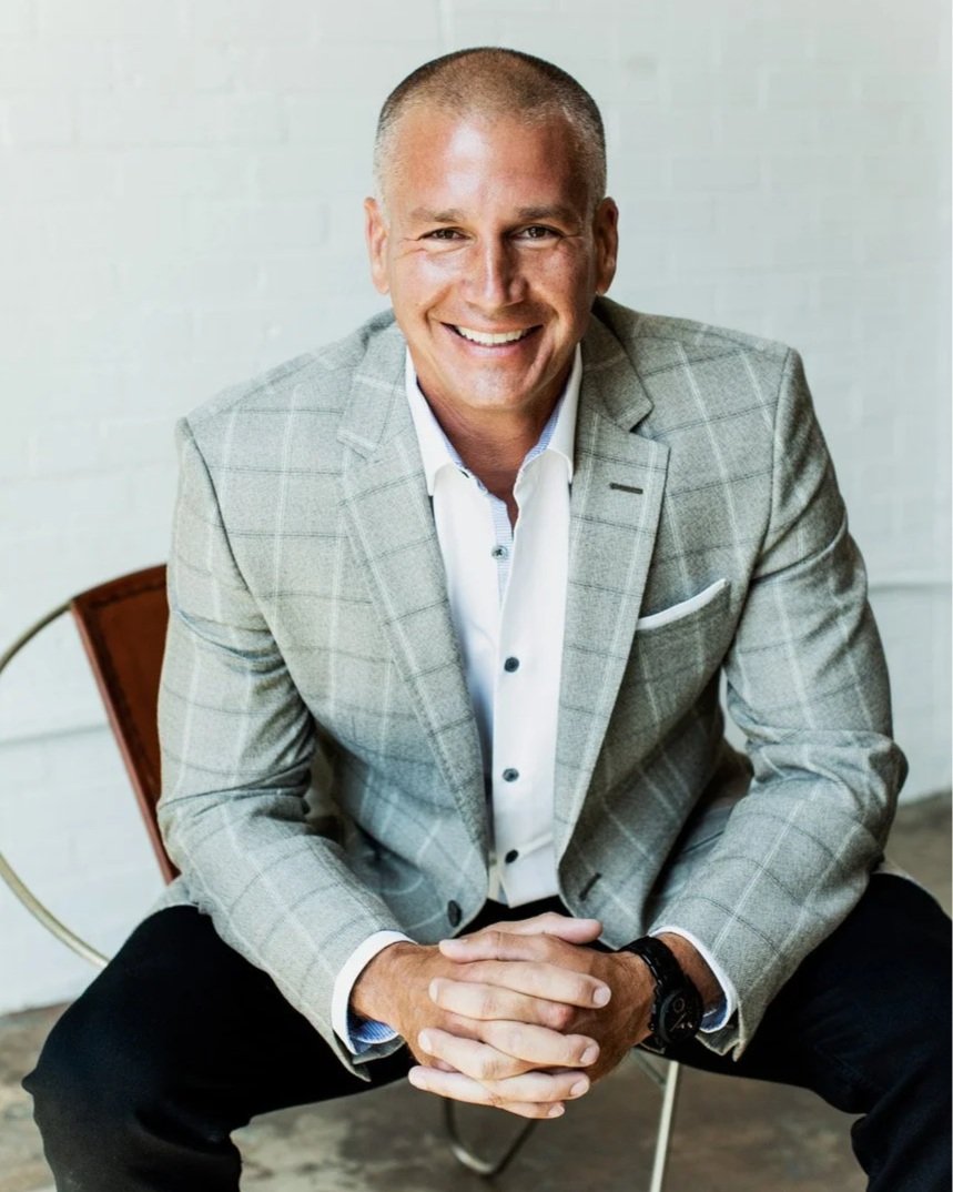 A smiling man in a gray checkered blazer and white shirt, sitting on a chair against a white brick wall.