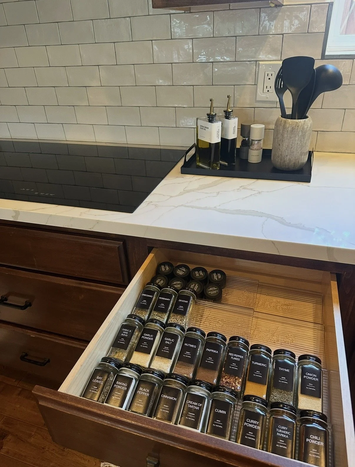 Open kitchen drawer filled with various spices in glass jars with black lids, labeled with different seasonings.