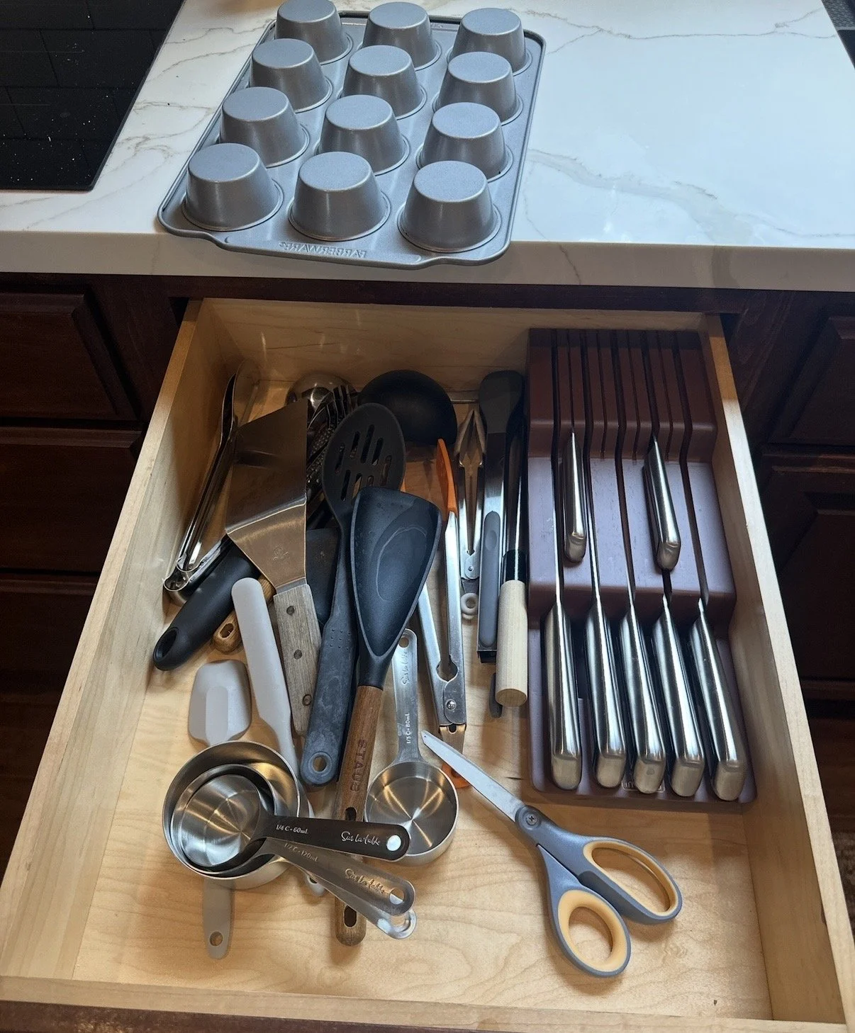 Open kitchen drawer containing various cooking utensils, including metal measuring cups, scissors, a vegetable peeler, spatulas, tongs, and a small silicone spatula.