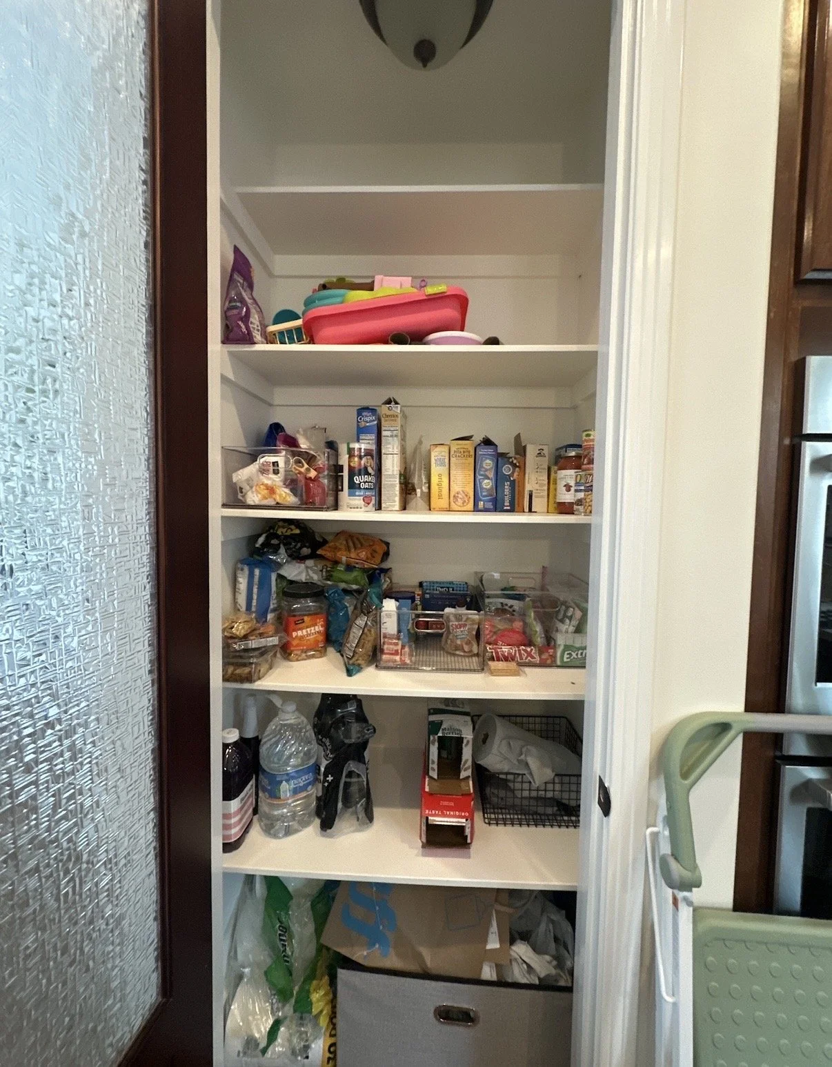 A pantry with five shelves containing snacks, canned goods, and miscellaneous household items. The bottom shelf has bags and a storage bin.