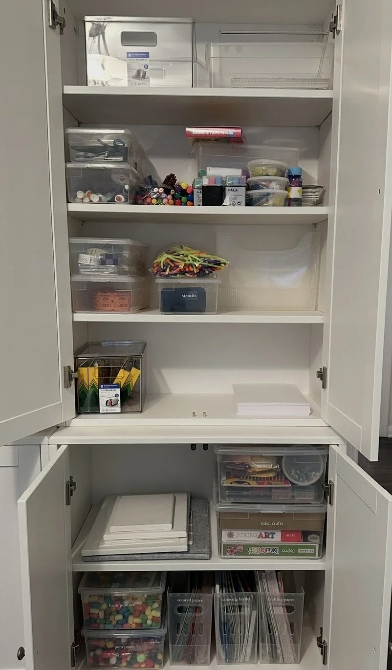 An organized white storage cabinet with three shelves and two lower compartments, containing craft supplies such as markers, scissors, glue, foam shapes, colored paper, and storage boxes.