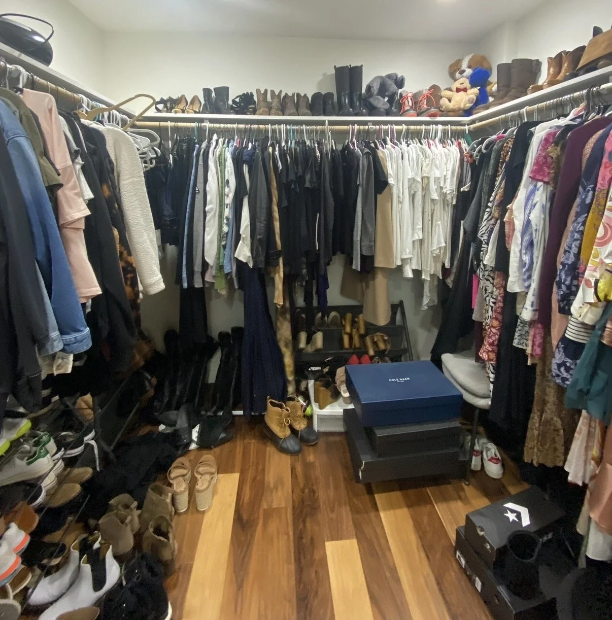 A walk-in closet filled with hanging clothes, shoes on shelves and the floor, and accessories on a shoe rack, with some boxes on the floor.