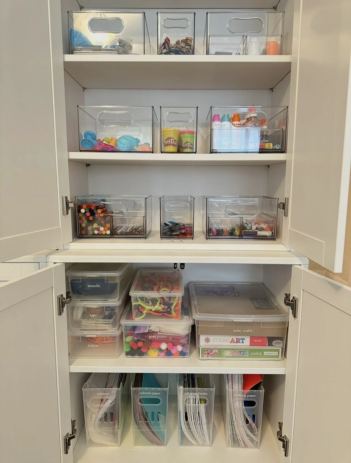 Organized white storage cabinet with craft supplies, including containers of markers, colored pencils, scissors, glue, tape, paint, and paper.
