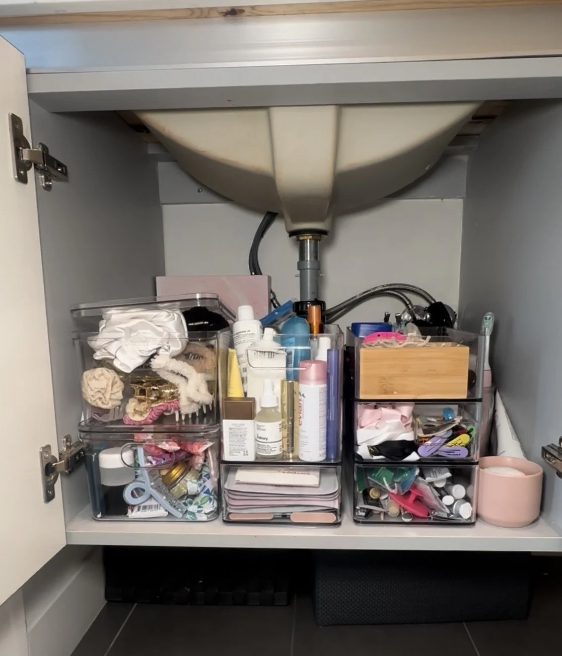 Under-sink cabinet with various makeup, skincare, and cosmetic items in clear storage containers.